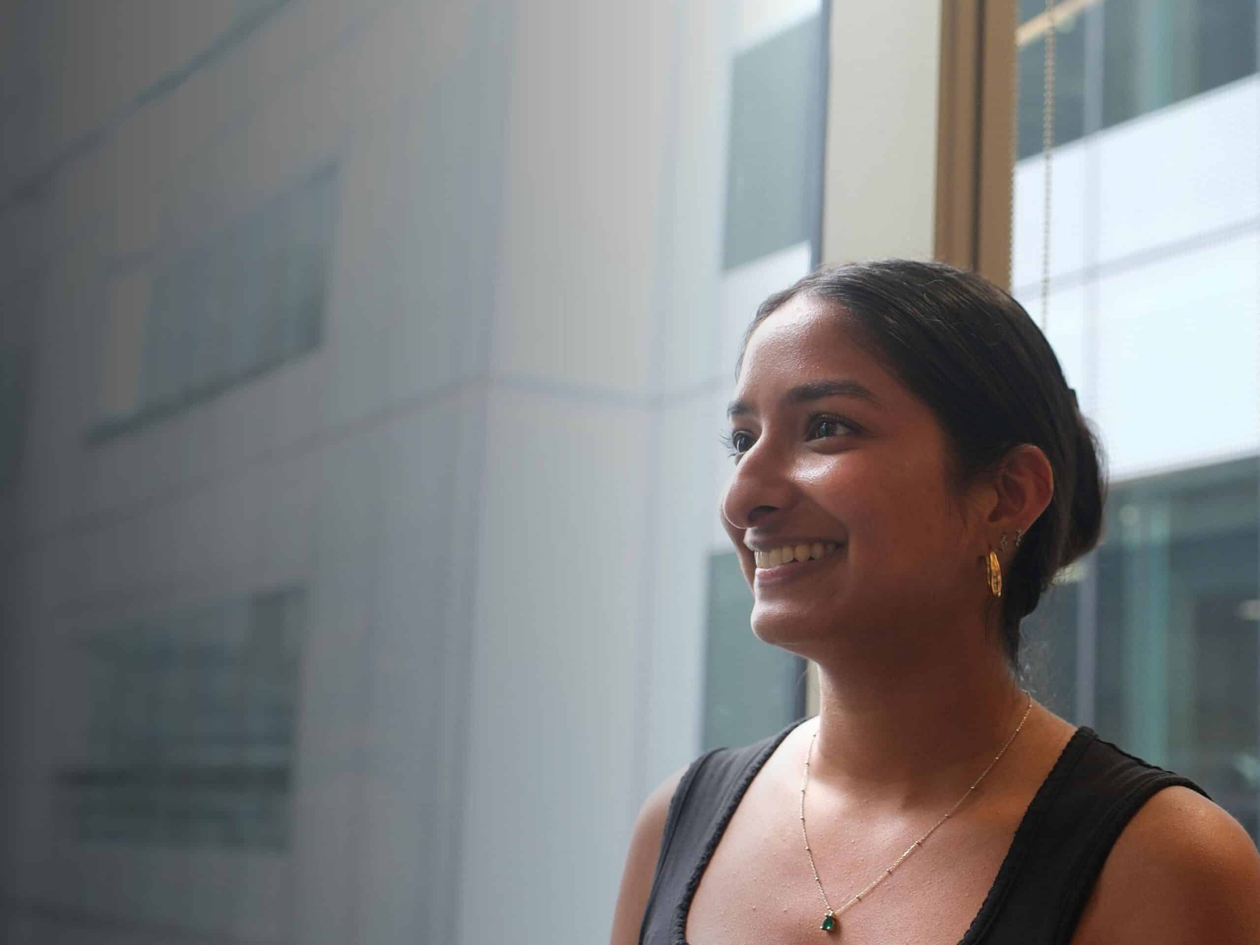 A young woman with dark hair smiles broadly, looking to the right. She wears a black top, gold hoop earrings, and a gold necklace with a green pendant.