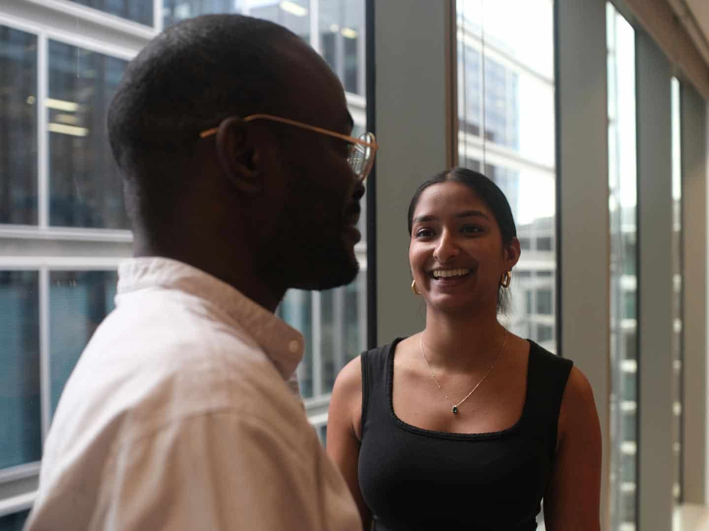 A man's back of head and side profile in the foreground, facing a smiling woman in the background. Both are indoors near large windows.