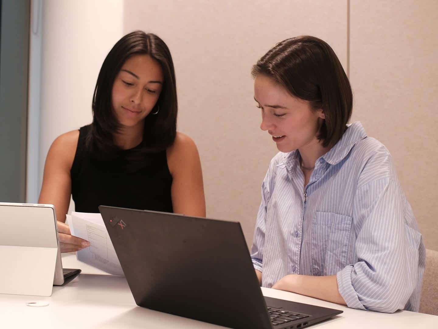 Two women at a table, one holding papers and the other looking at a laptop.