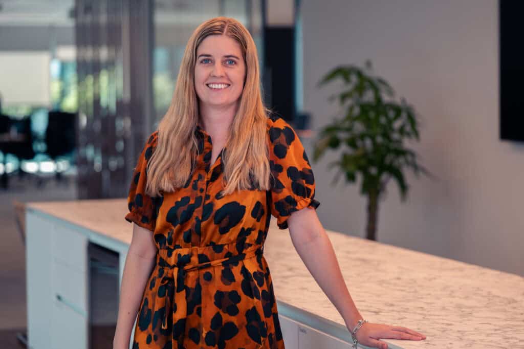 A smiling woman with long blonde hair in an orange and black leopard print dress, standing next to a marble-patterned counter in an office setting.