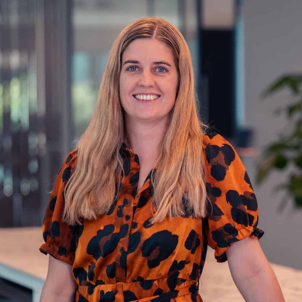 A smiling woman with long blonde hair in an orange and black leopard print dress, standing next to a marble-patterned counter in an office setting.