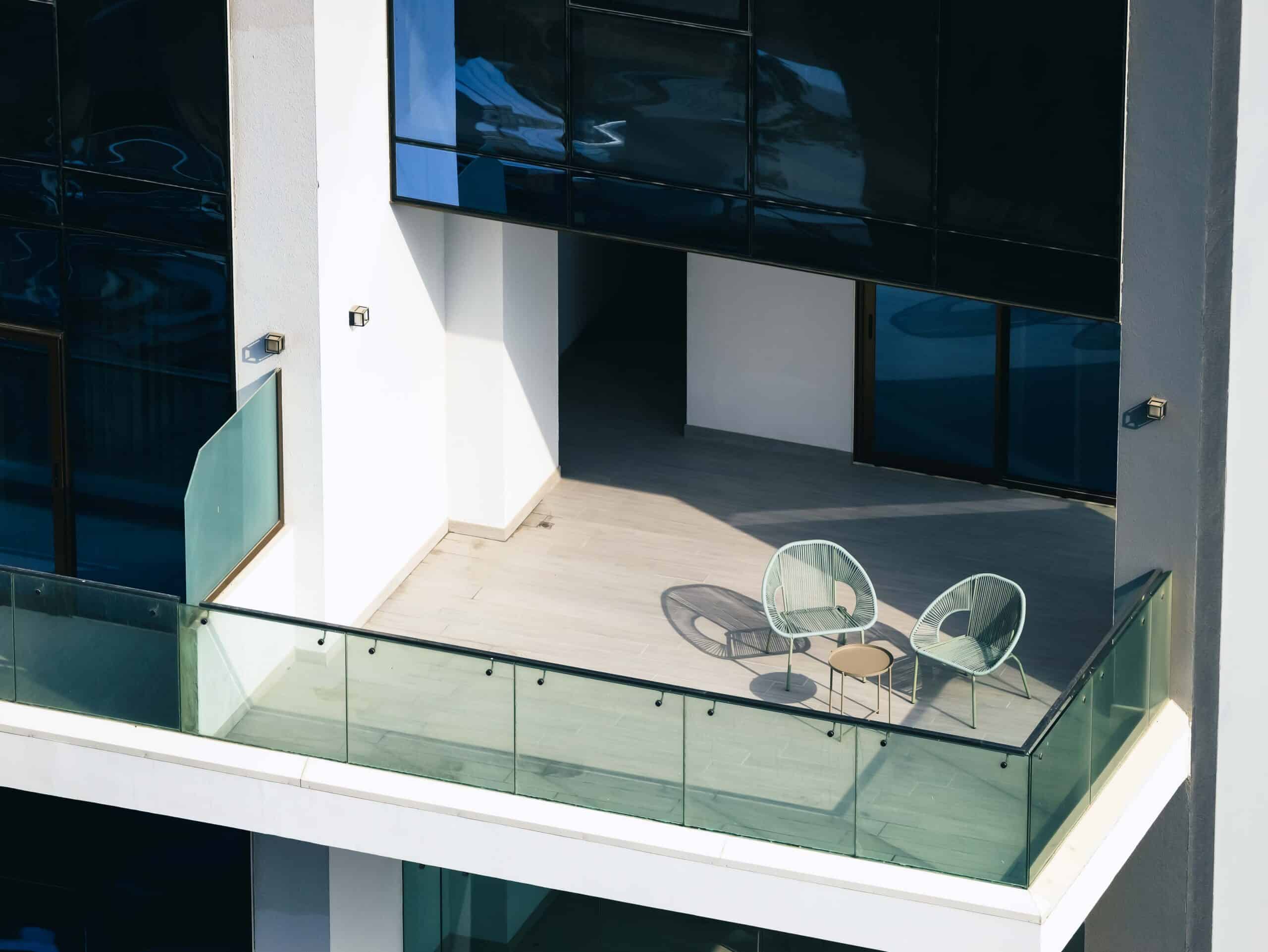 Modern balcony with two light green chairs, a small round table, glass railing, and strong shadows on a light-colored tiled floor.