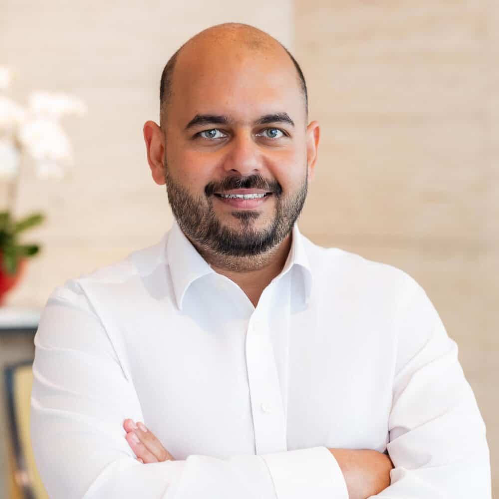 A man with a bald head and beard, wearing a white shirt, smiling with arms crossed.