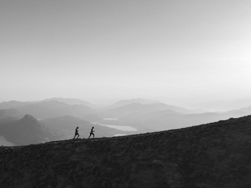 Black and white image of two people running on a grassy ridge with layered mountains and lakes in the background under a bright, hazy sky.