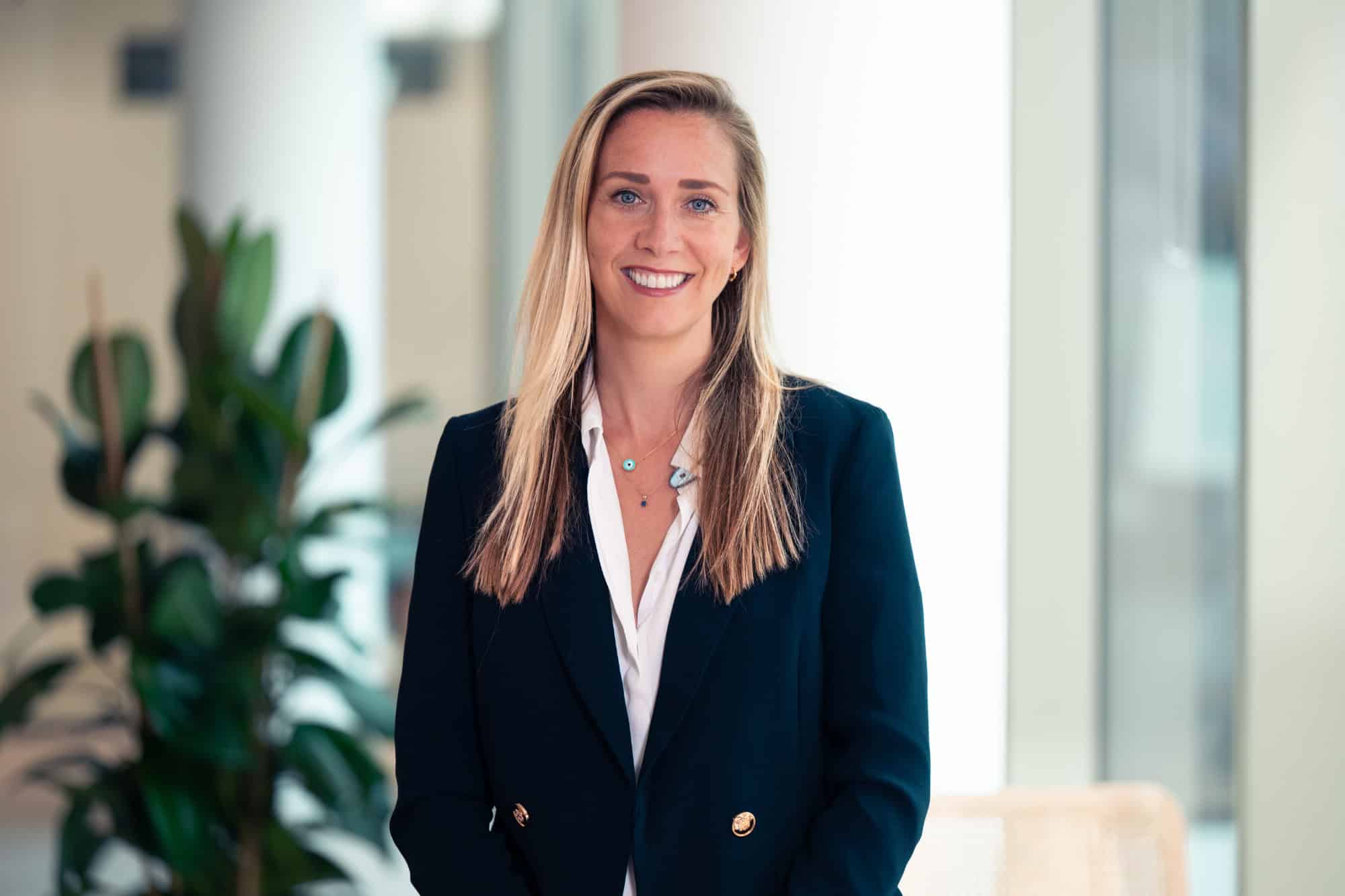 Headshot of a smiling woman with long blonde hair, blue eyes, wearing a dark blue blazer and white shirt.