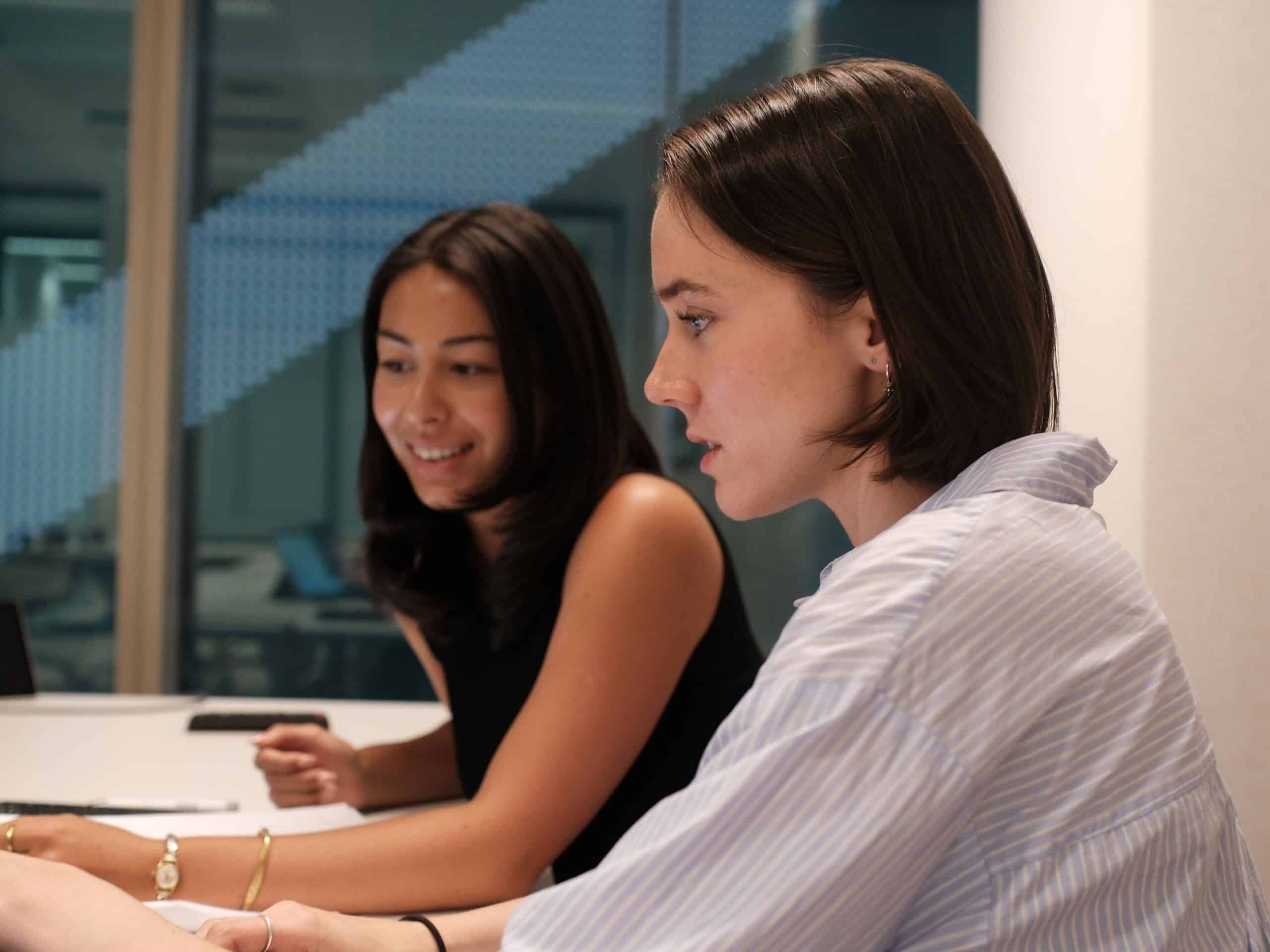 Two young women, one in a striped shirt and one in a black top, seated at a table, engaged in discussion.