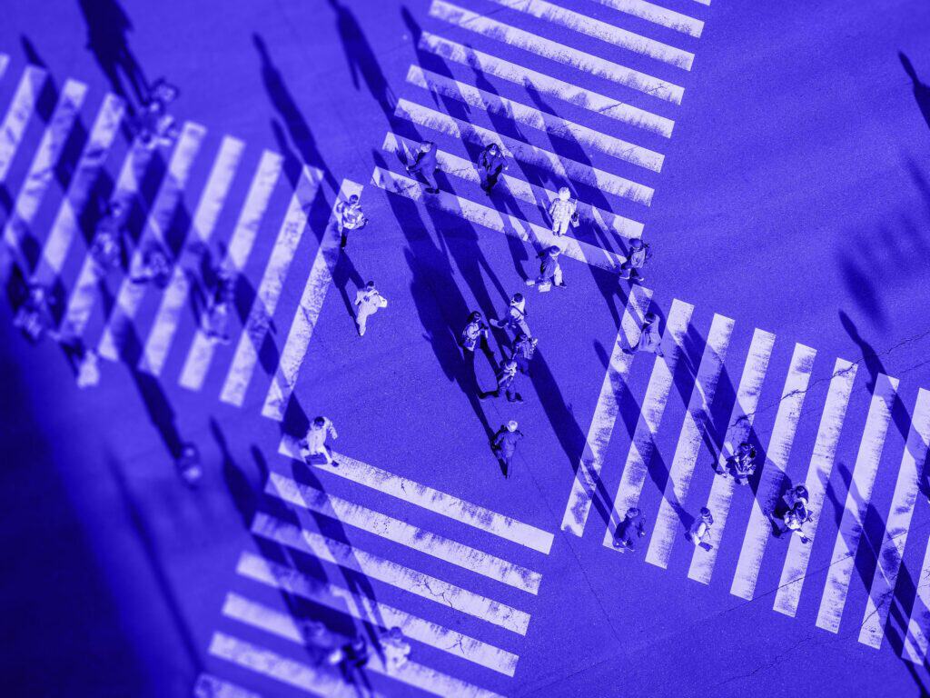 Aerial view of multiple people crossing a street at a crosswalk, rendered in shades of blue with long shadows.