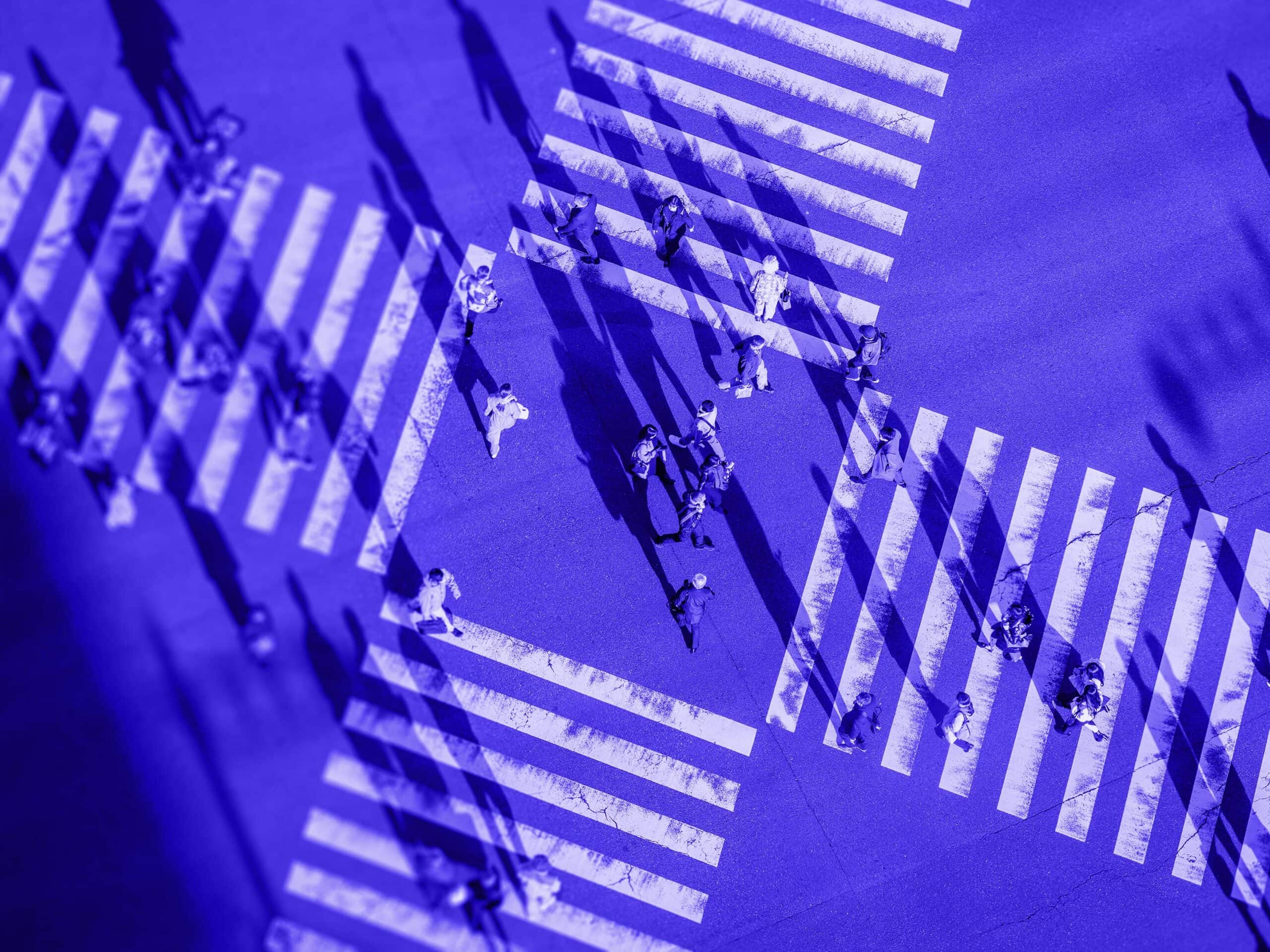 Aerial view of multiple people crossing a street at a crosswalk, rendered in shades of blue with long shadows.