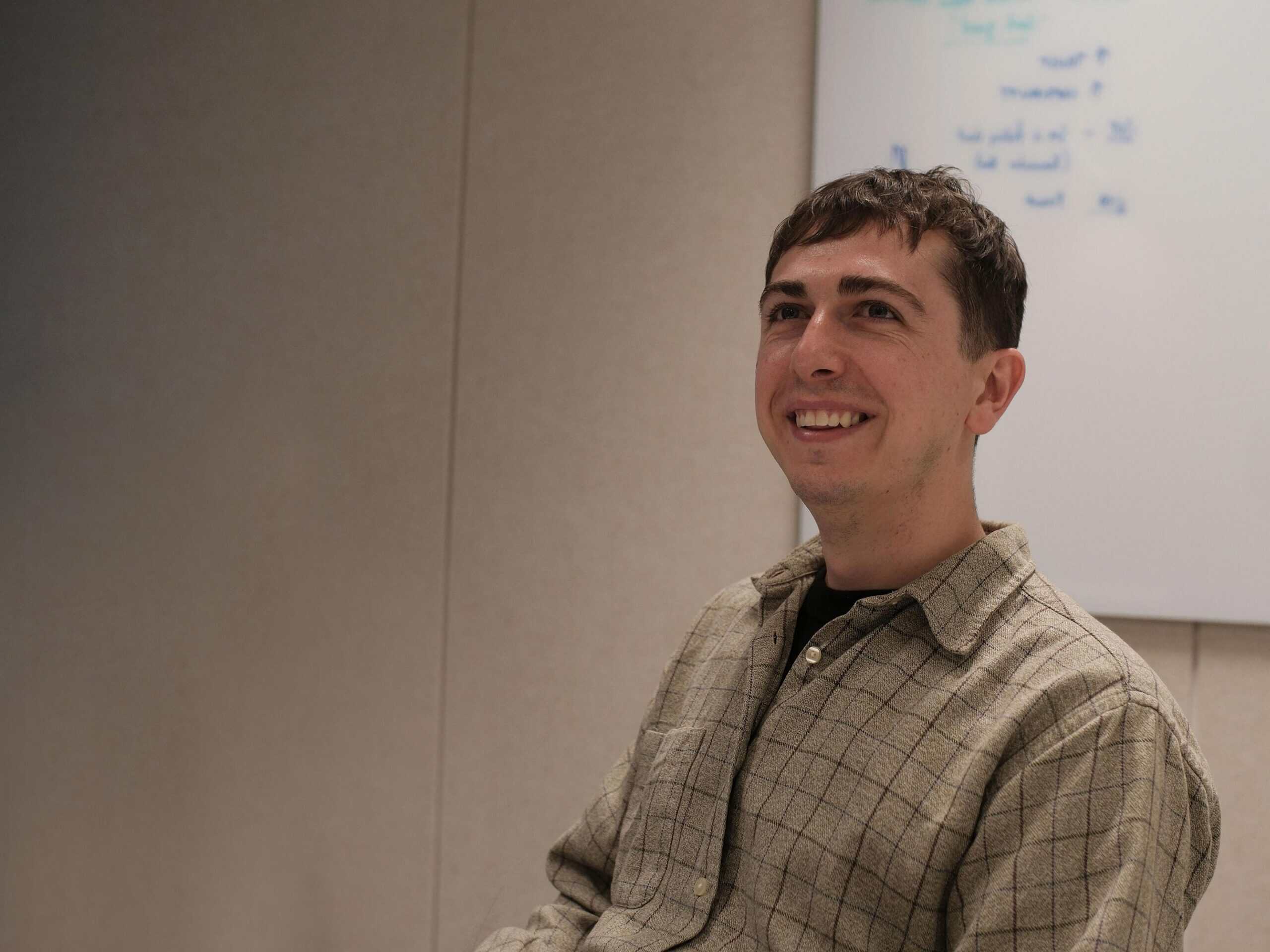 A man with short brown hair and a plaid shirt smiles, looking upwards and to the right, with a whiteboard in the background.