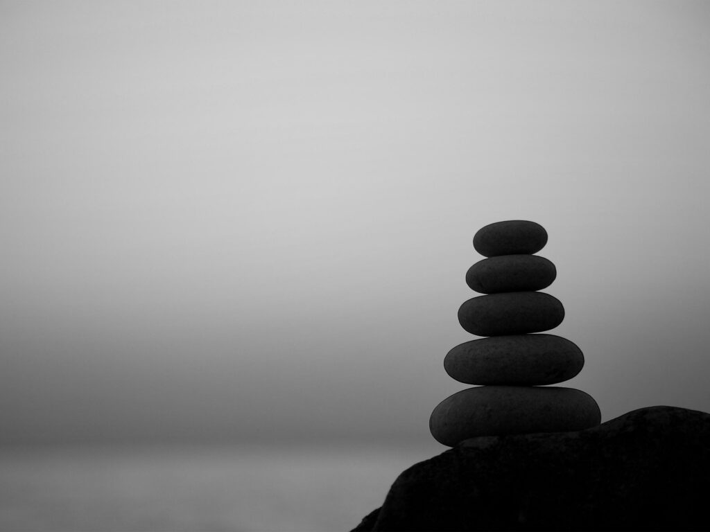 A black and white image of five smooth, dark stones stacked vertically on a larger rock, set against a light gray, hazy background.