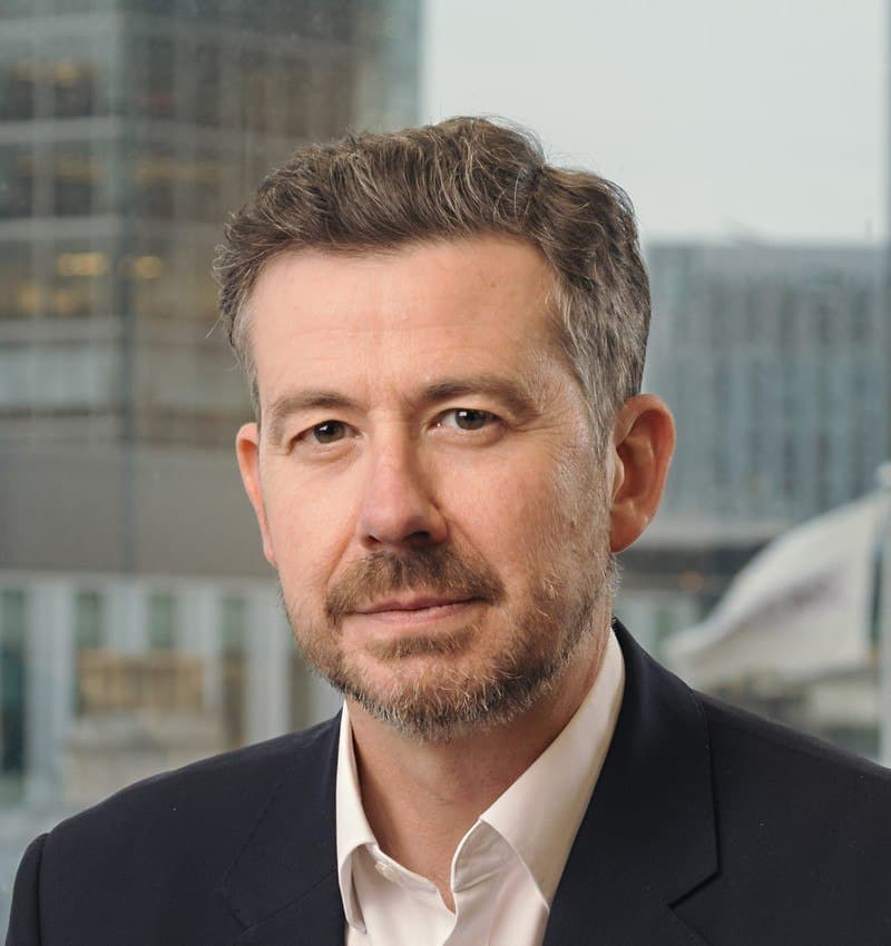 Headshot of a man with short, wavy hair and a beard, wearing a dark suit jacket and white shirt, looking forward.