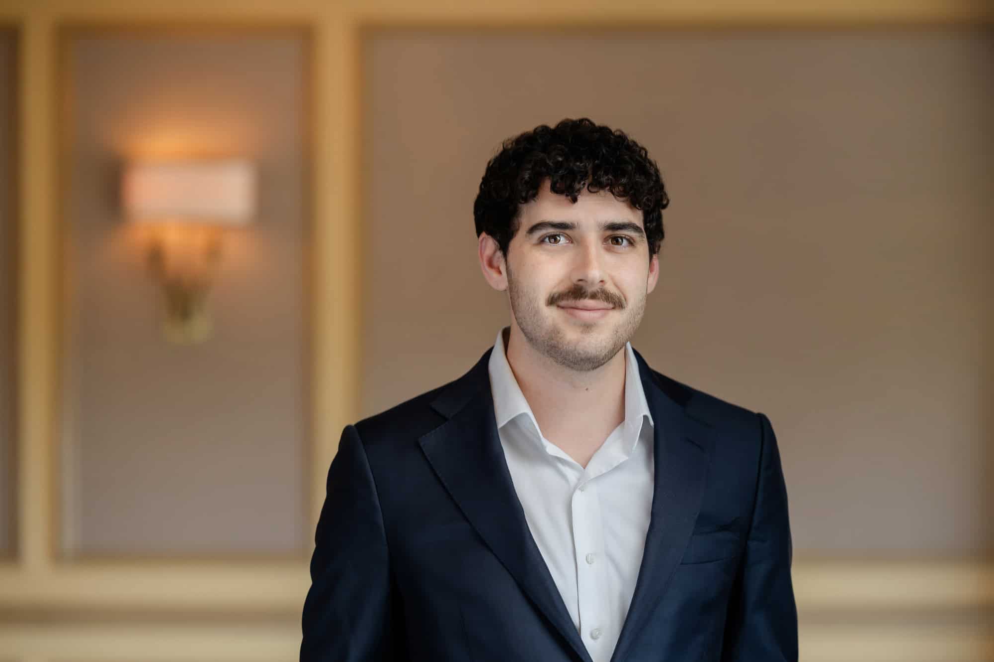 A man with dark curly hair and a mustache, wearing a dark blue suit and white shirt, smiles at the camera.