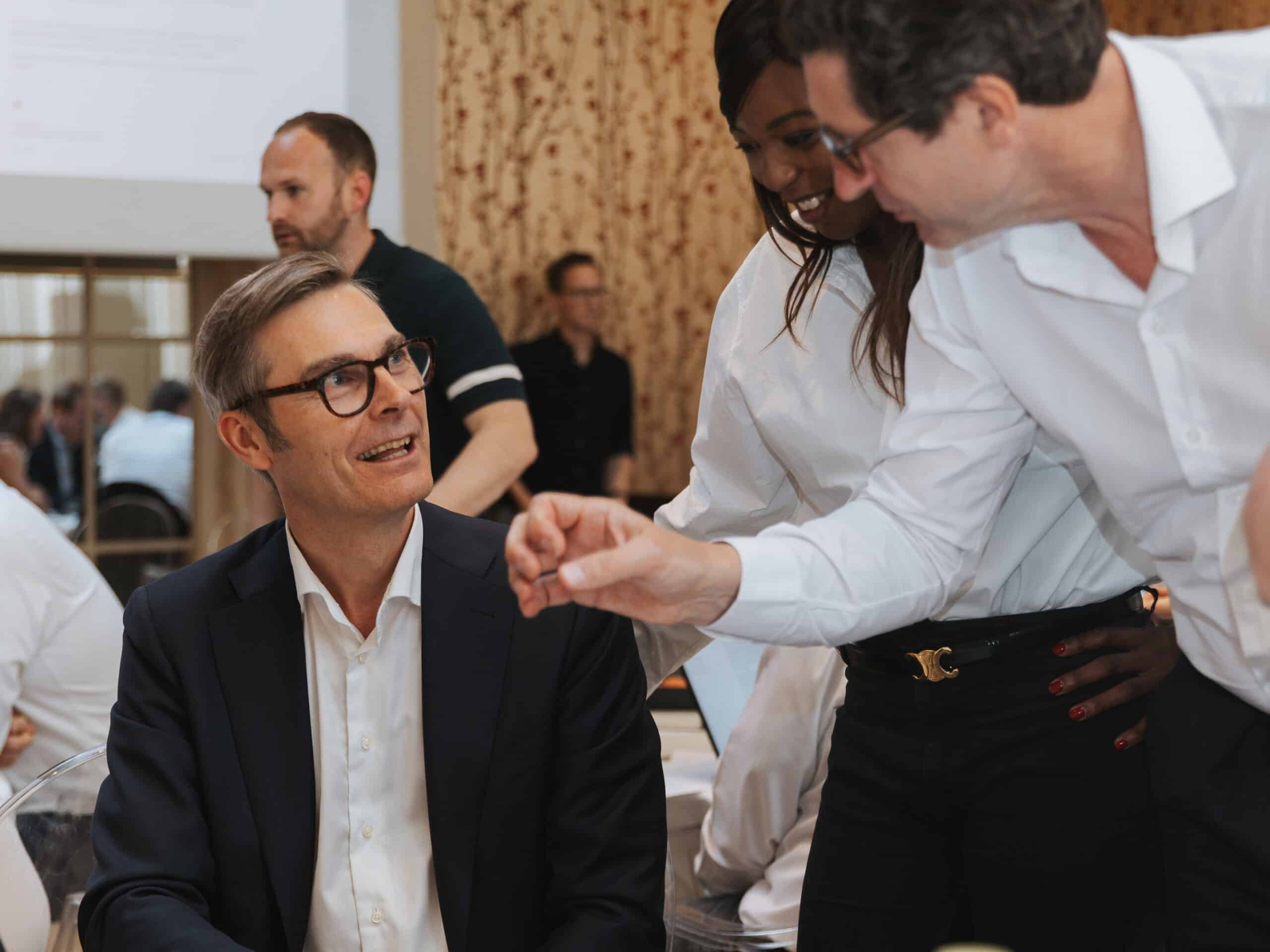 Three people, two men and one woman, are closely engaged in a discussion. One man is seated, smiling, while the woman and another man stand, leaning in and looking at something.