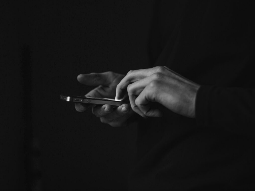 Black and white image of two hands holding and interacting with a glowing smartphone in darkness.