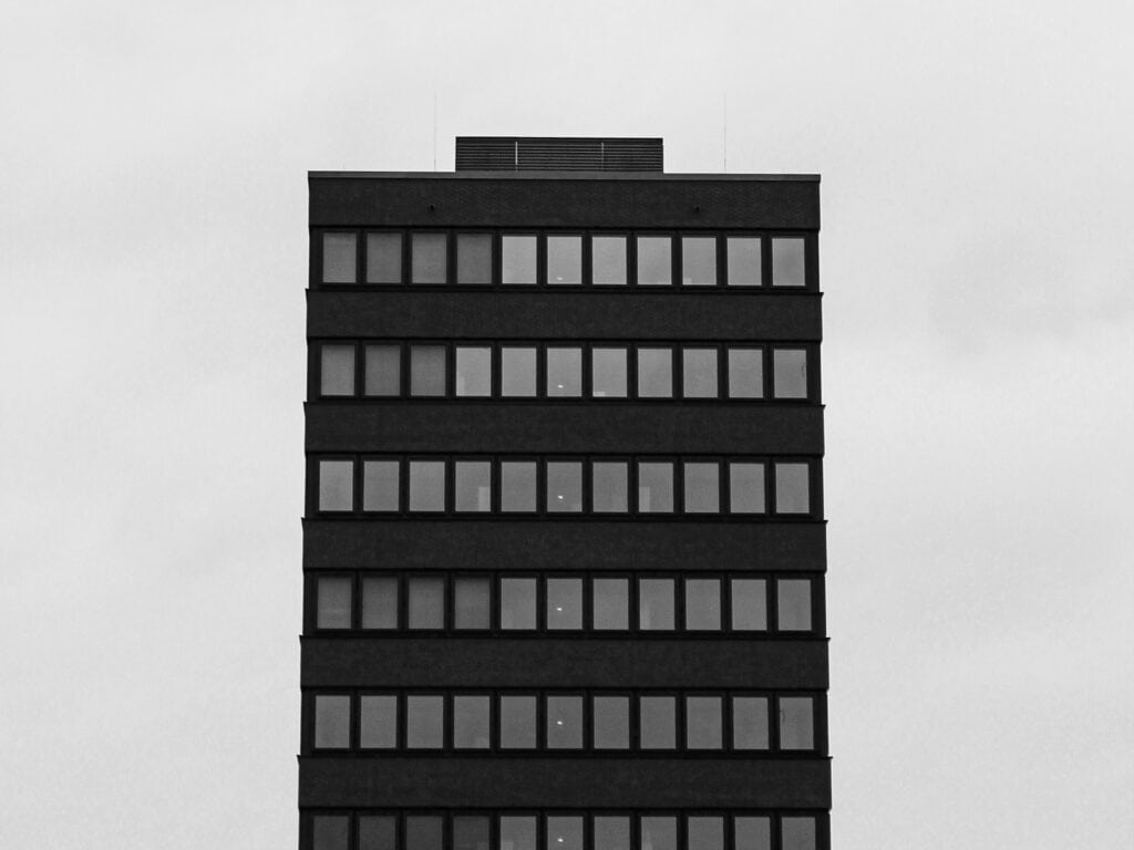 A tall, dark, rectangular building with many windows against a light gray sky, in black and white.