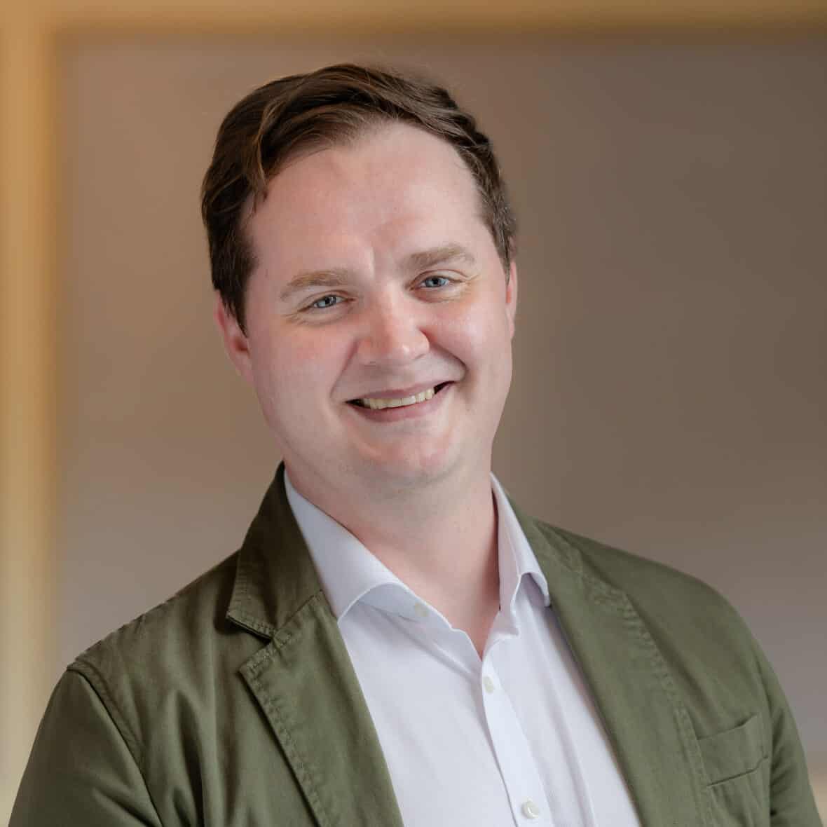 Headshot of a smiling man in a white shirt and olive green jacket.