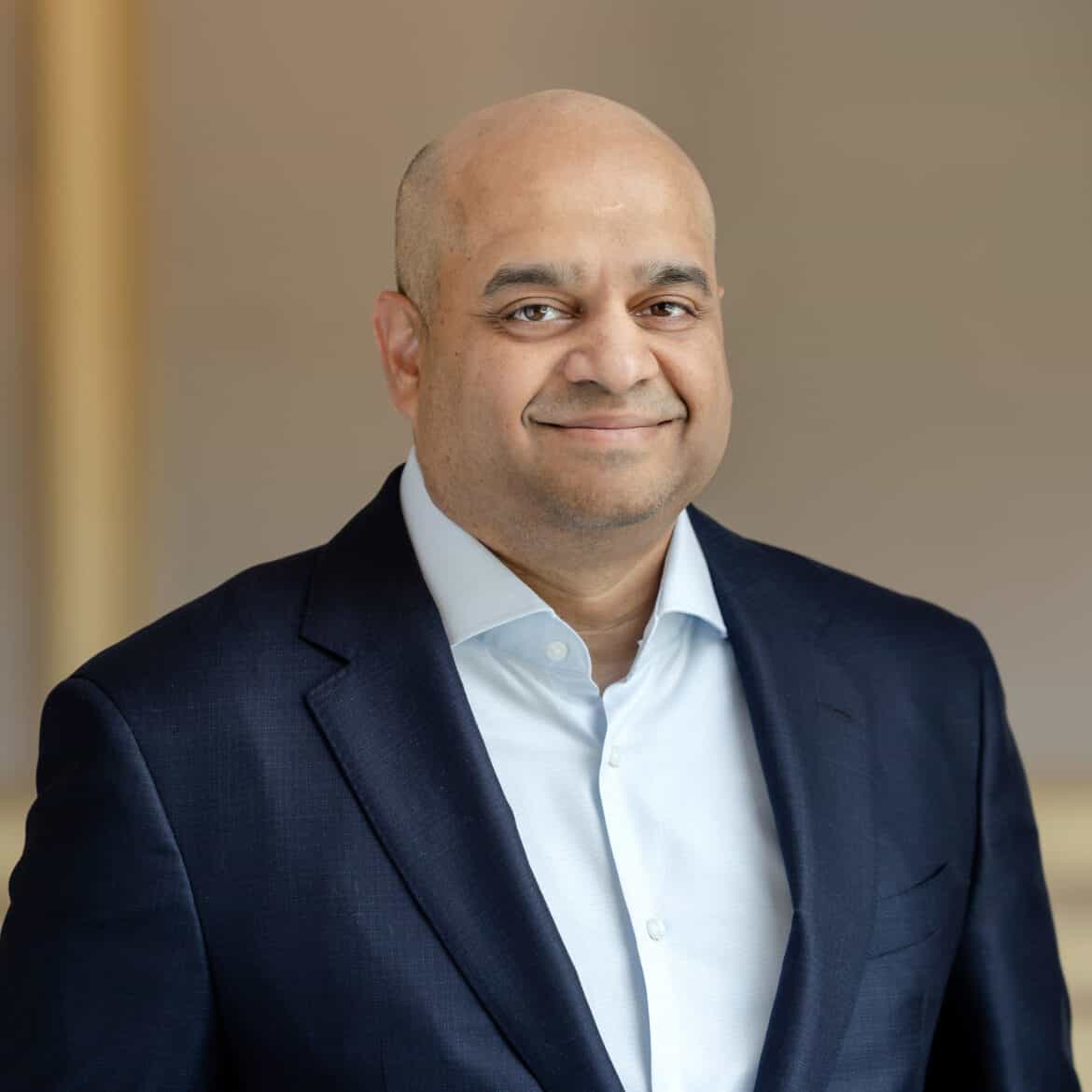 Headshot of a bald man with a light brown complexion, smiling, wearing a dark blue suit jacket and light blue shirt.