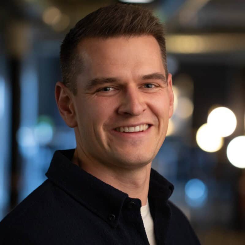Headshot of a smiling man with short brown hair and blue eyes, wearing a dark blue shirt over a light t-shirt. Blurred lights are in the background.