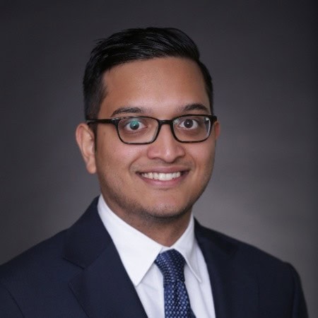 Headshot of a smiling man with dark hair and glasses, wearing a navy suit and blue tie.