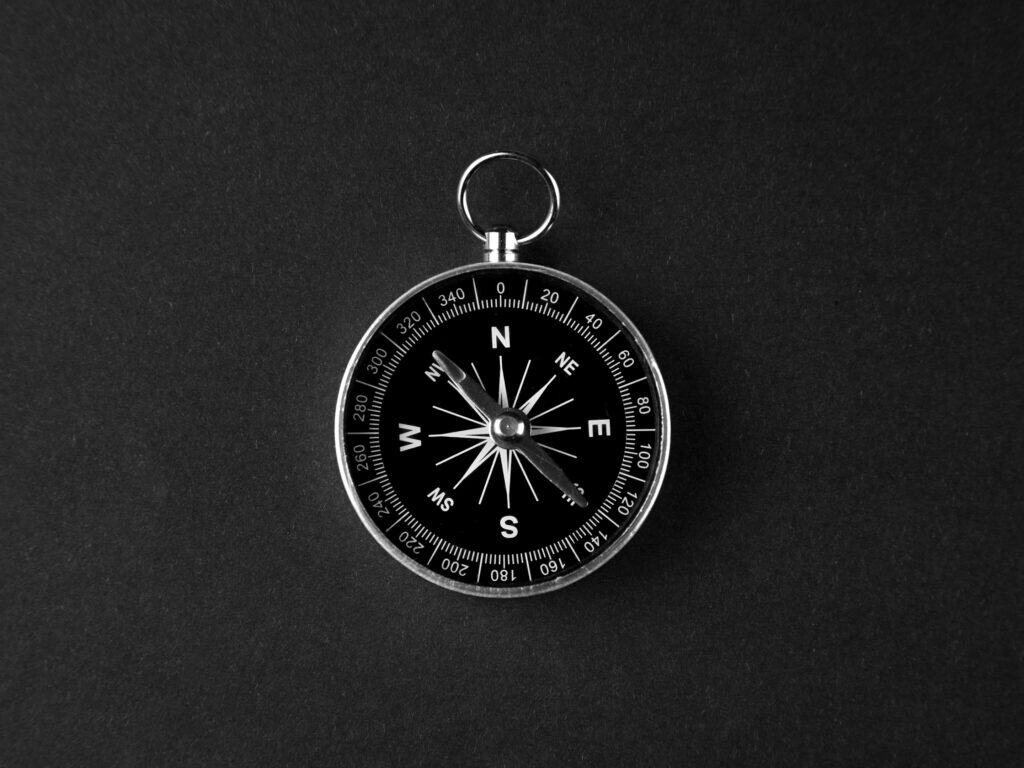 A black and white image of a compass with a silver casing and a black face, showing cardinal directions and a needle pointing North, on a dark background.