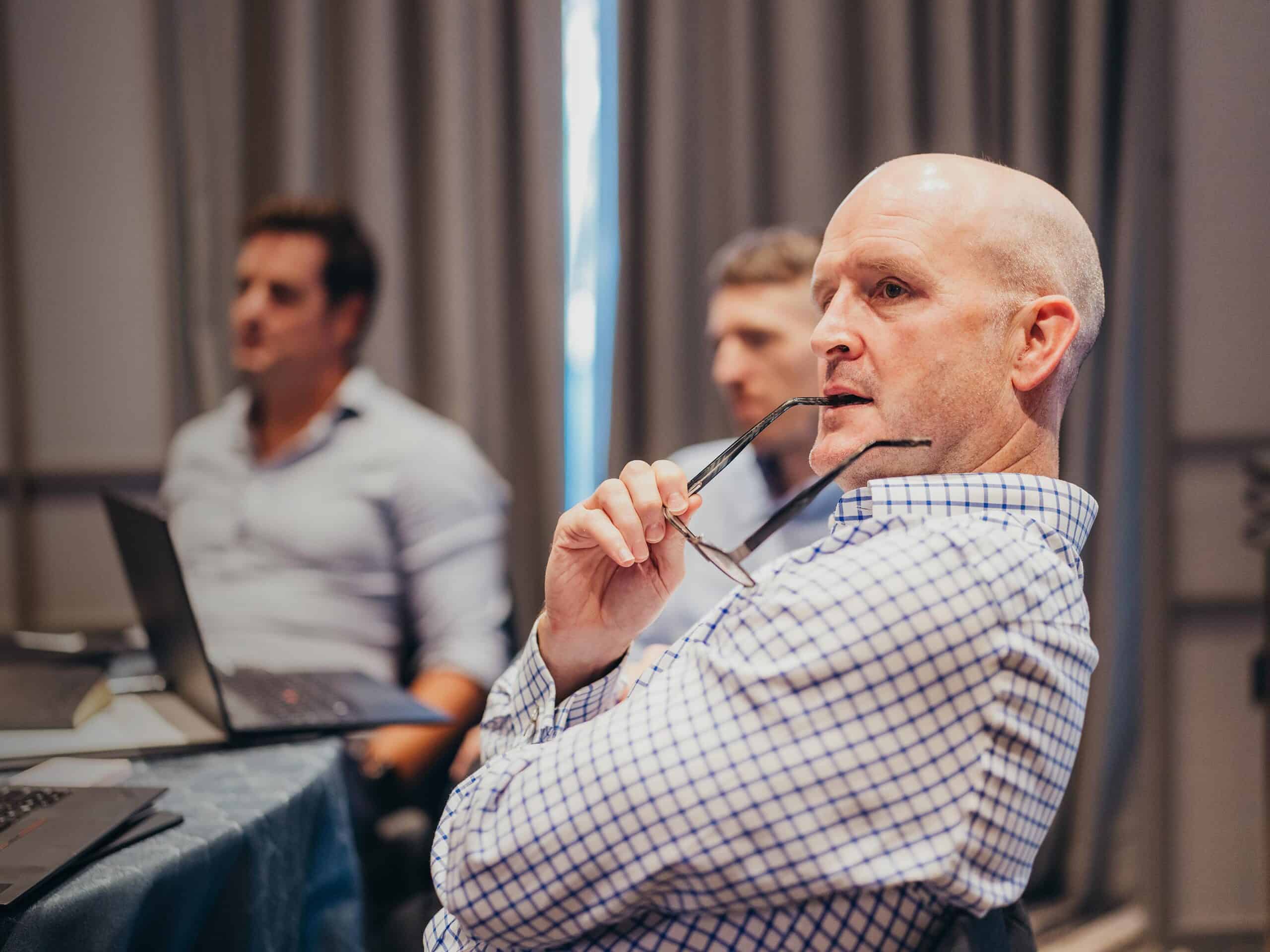 Side profile of a bald man in a plaid shirt, holding eyeglasses to his mouth, listening in a meeting.