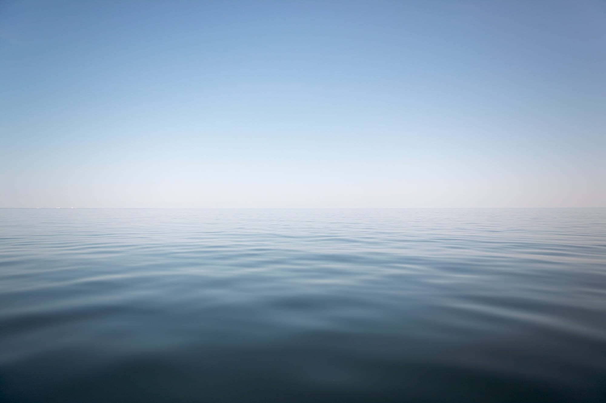 A minimalist image of calm, blue water meeting a clear, light blue sky at a sharp horizon line.