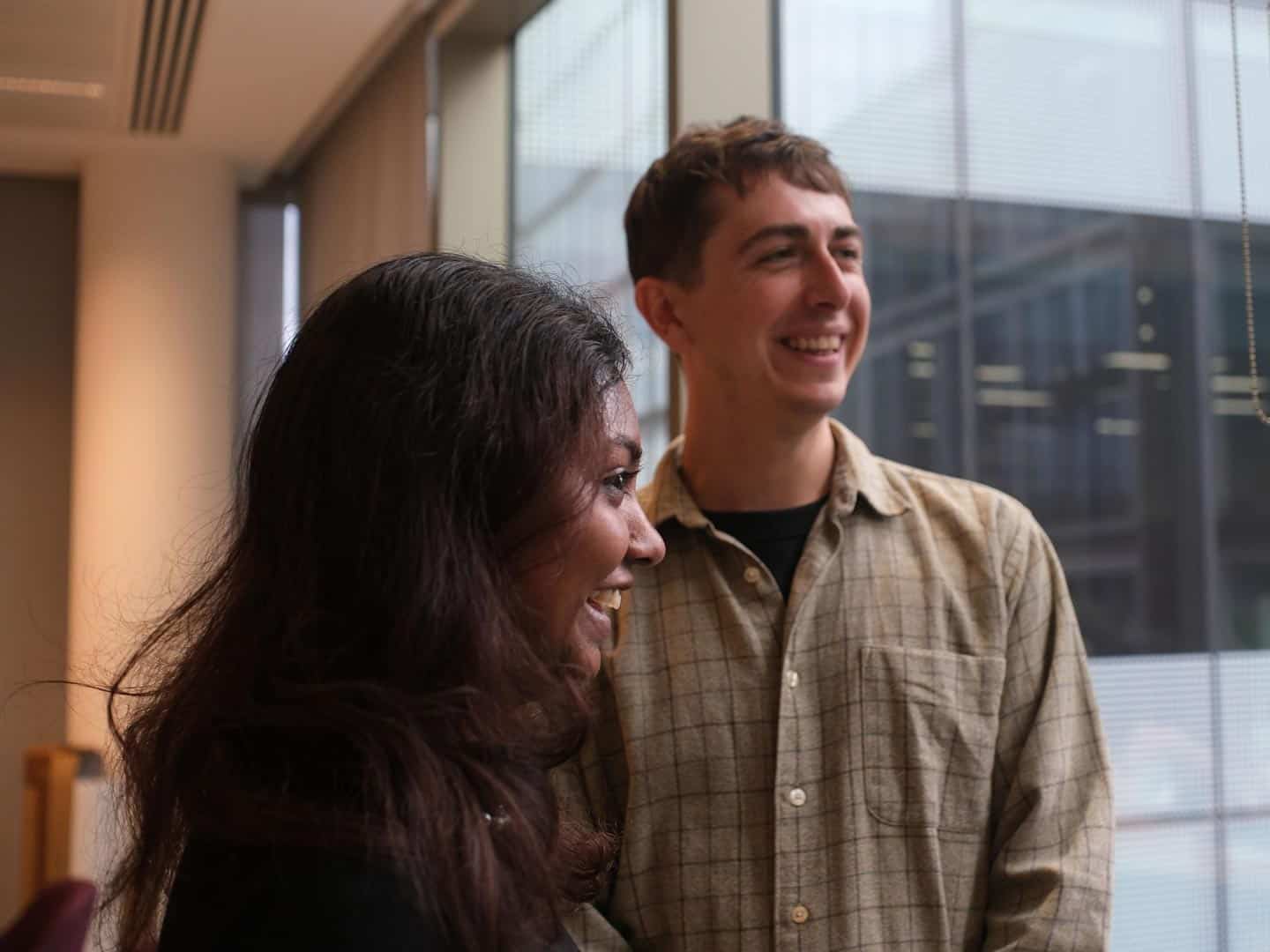 A woman and a man, both smiling, look towards the right. The woman has long dark hair, and the man wears a plaid shirt, with a window and buildings in the background.