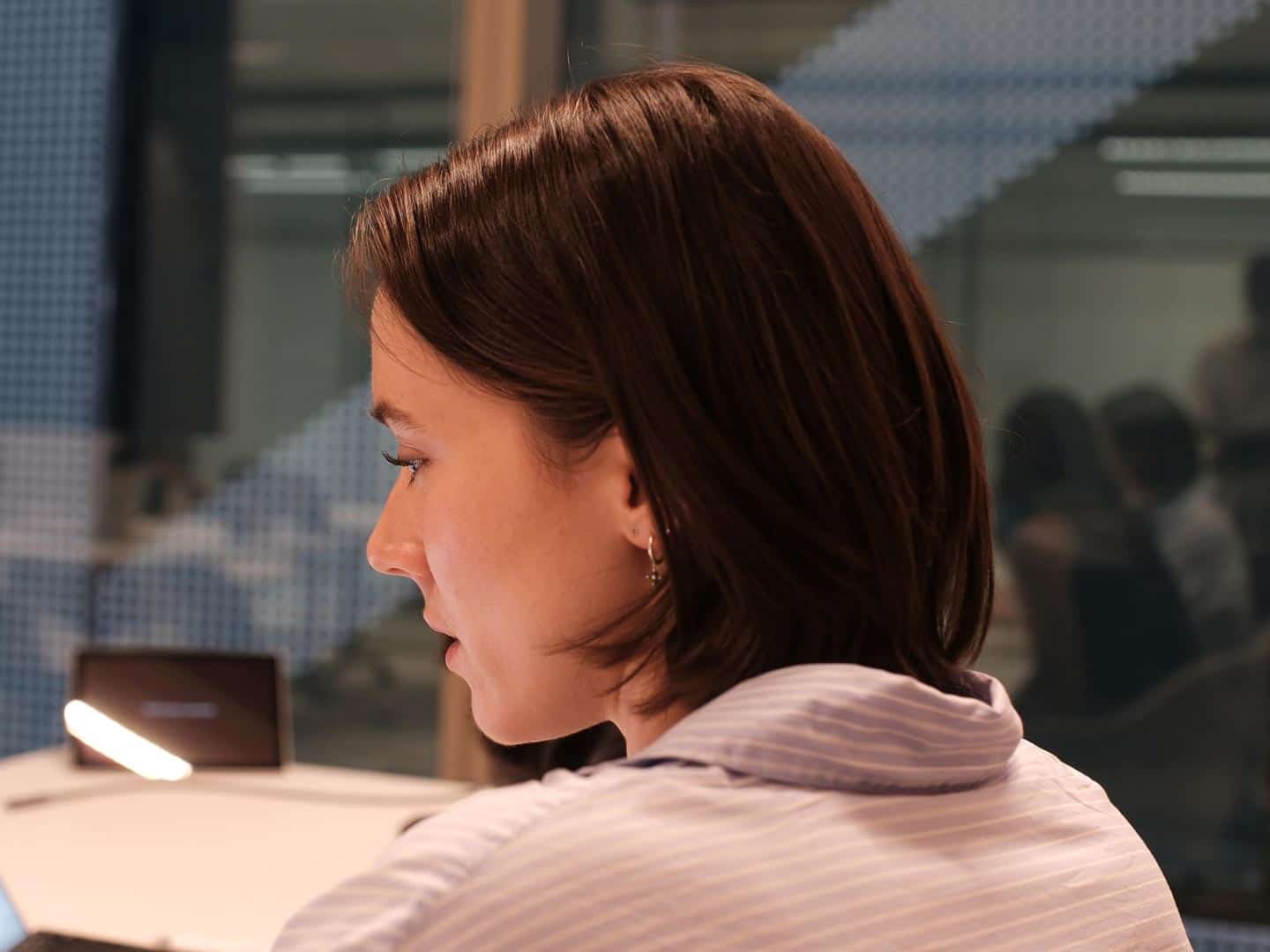 Profile view of a woman with short brown hair and a striped shirt, looking down at a desk with a bright lamp. A blurred office background is visible through a glass partition.