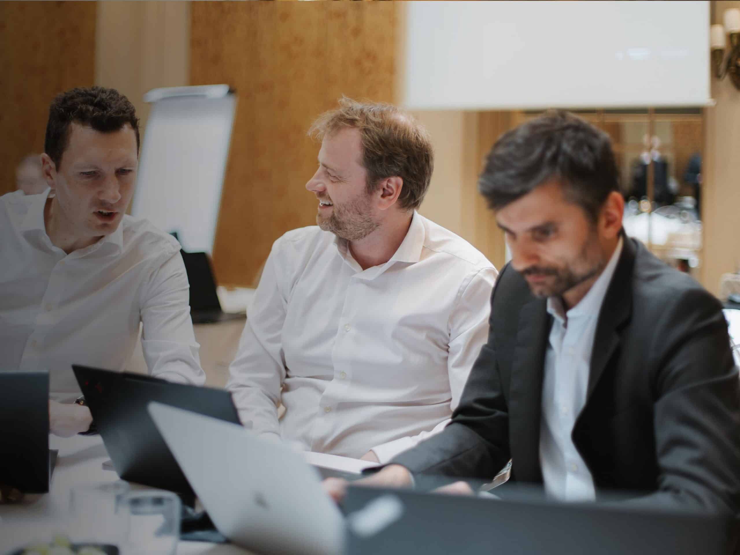 Three men in business attire, two in white shirts and one in a suit, are seated at a table with laptops, engaged in a meeting. The man in the middle is smiling and looking at the man on the left.