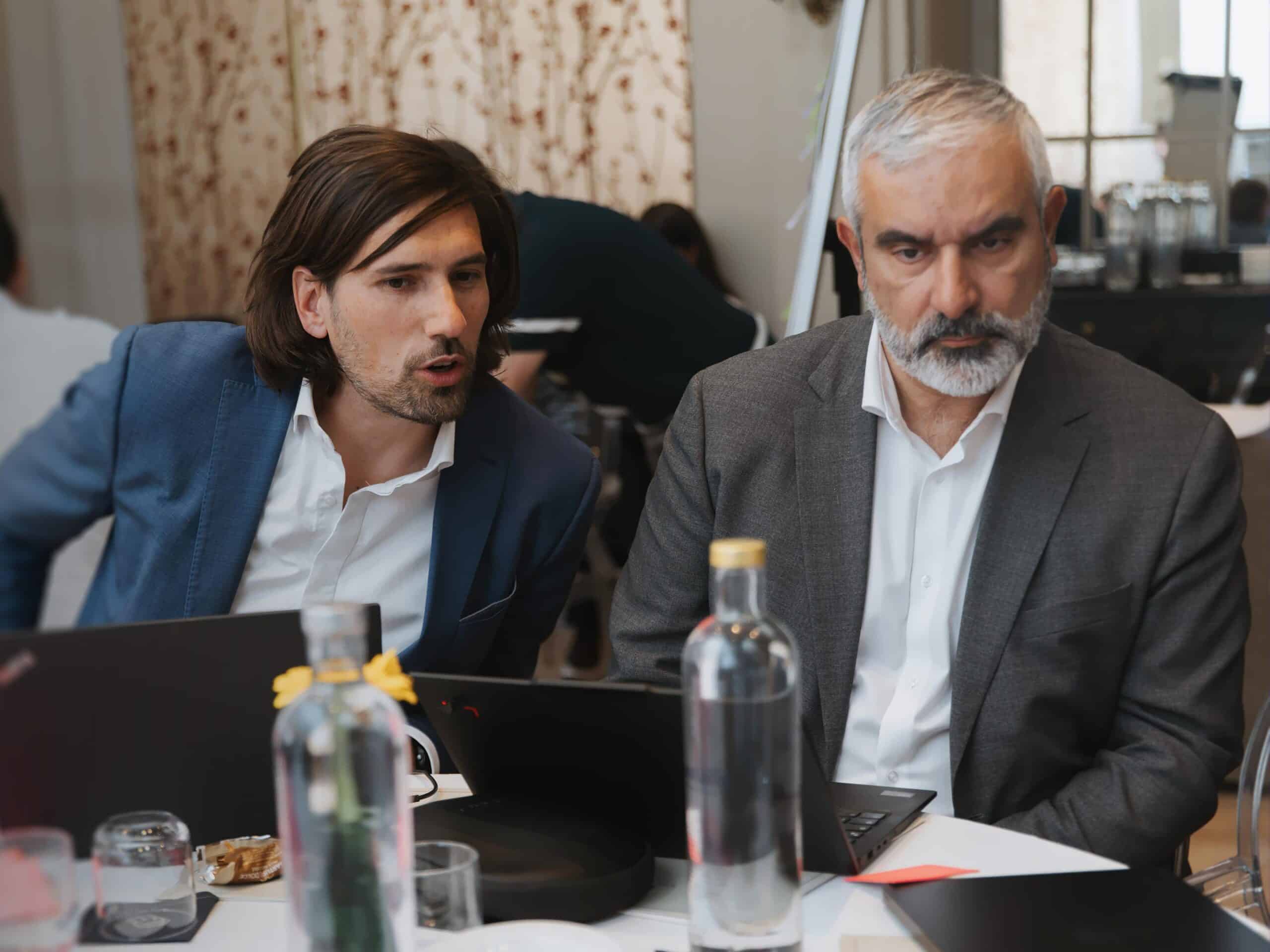 Two men in business attire sit at a table with laptops and water bottles. One man with long dark hair speaks, while the other with grey hair and beard looks down.