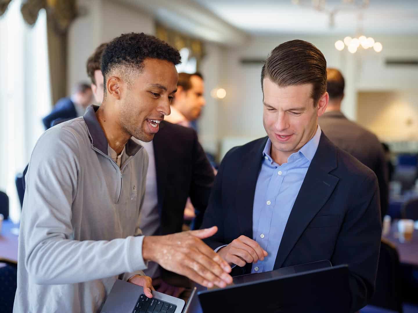 Two men, one in a grey pullover and one in a suit jacket, looking at a laptop screen and discussing its content.