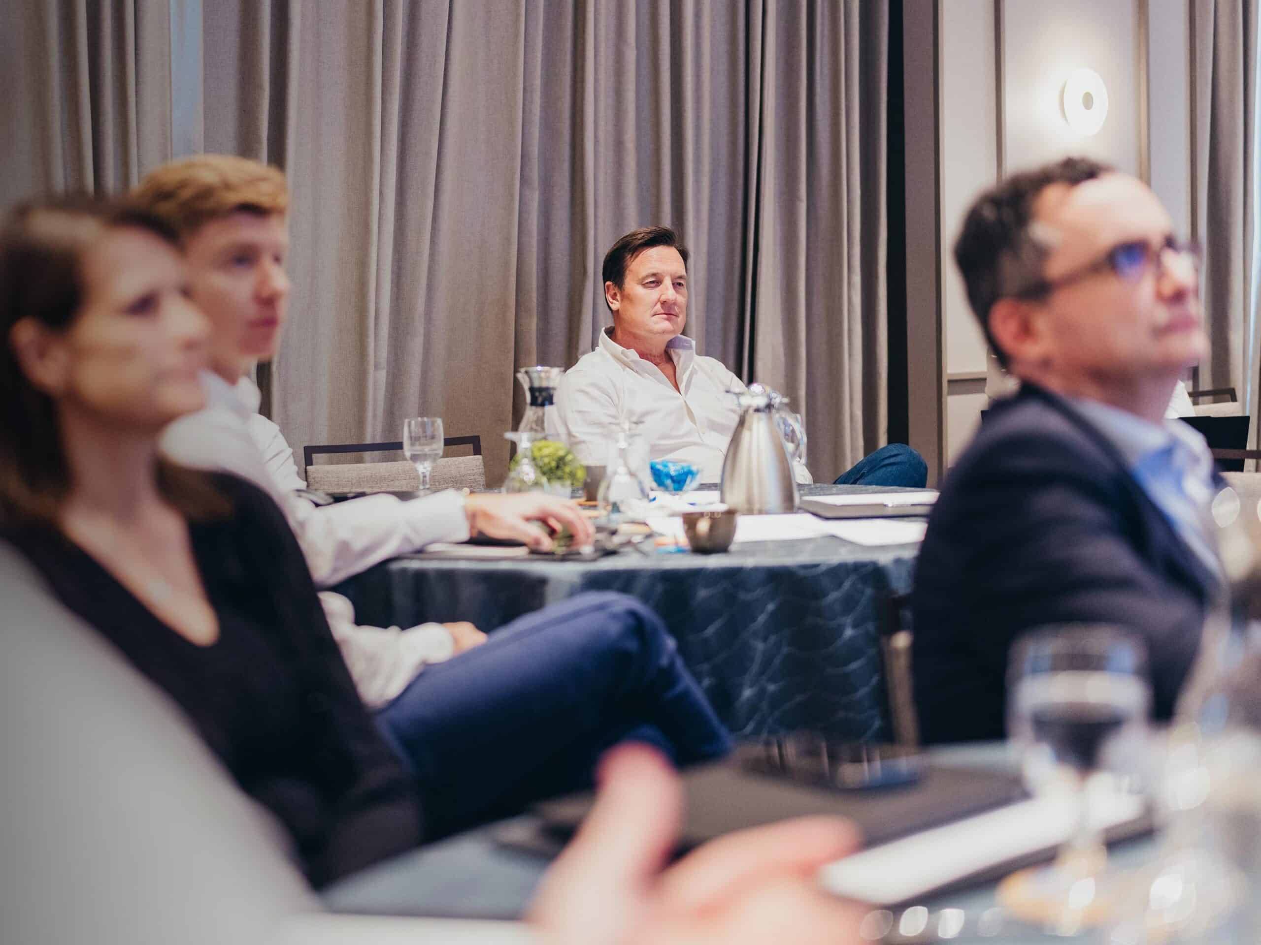 Four people seated at a table in a meeting room, with one man in a white shirt in focus, looking to the right.