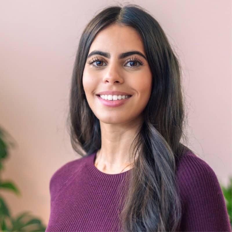 Headshot of a smiling woman with long dark hair wearing a plum sweater against a pink background.