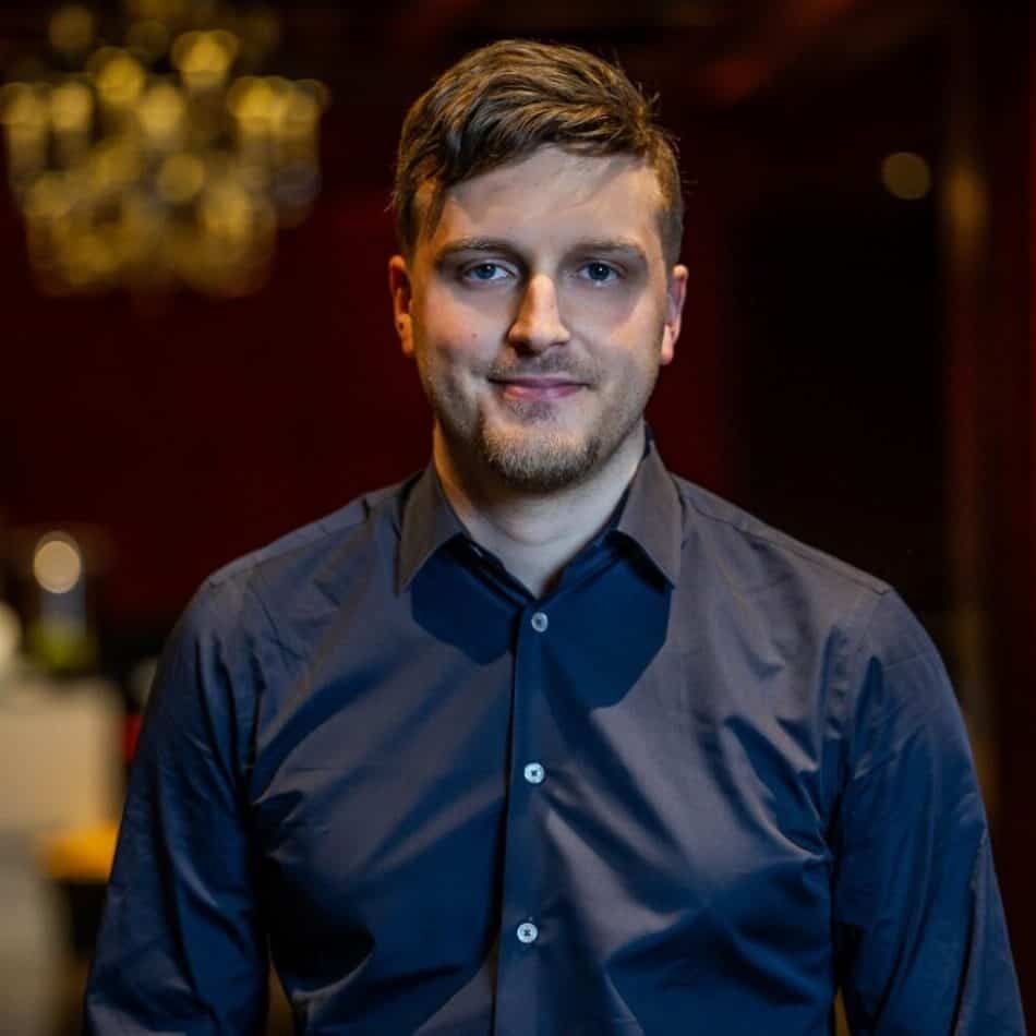 A man with short brown hair and a light beard smiles at the camera, wearing a dark blue shirt. The background is blurred with red and gold tones.