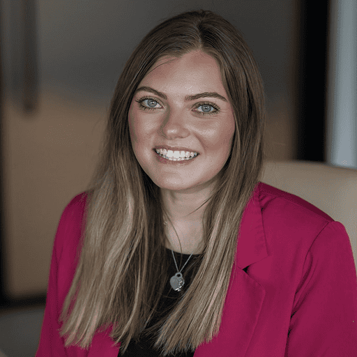 Headshot of a smiling young woman with light brown hair and blue-green eyes, wearing a magenta blazer and a heart necklace.