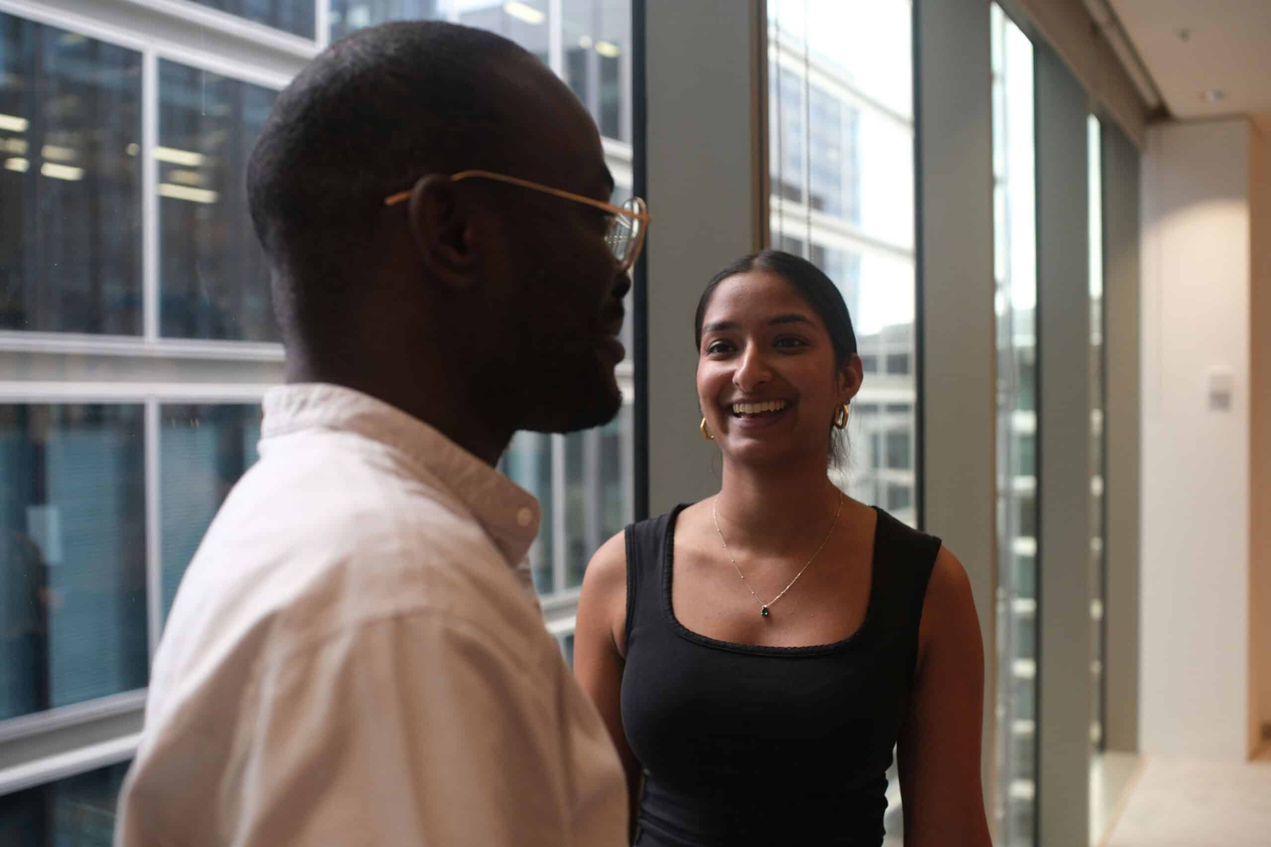 A man and a woman talking by large windows. The woman smiles at the man, who is seen from behind.