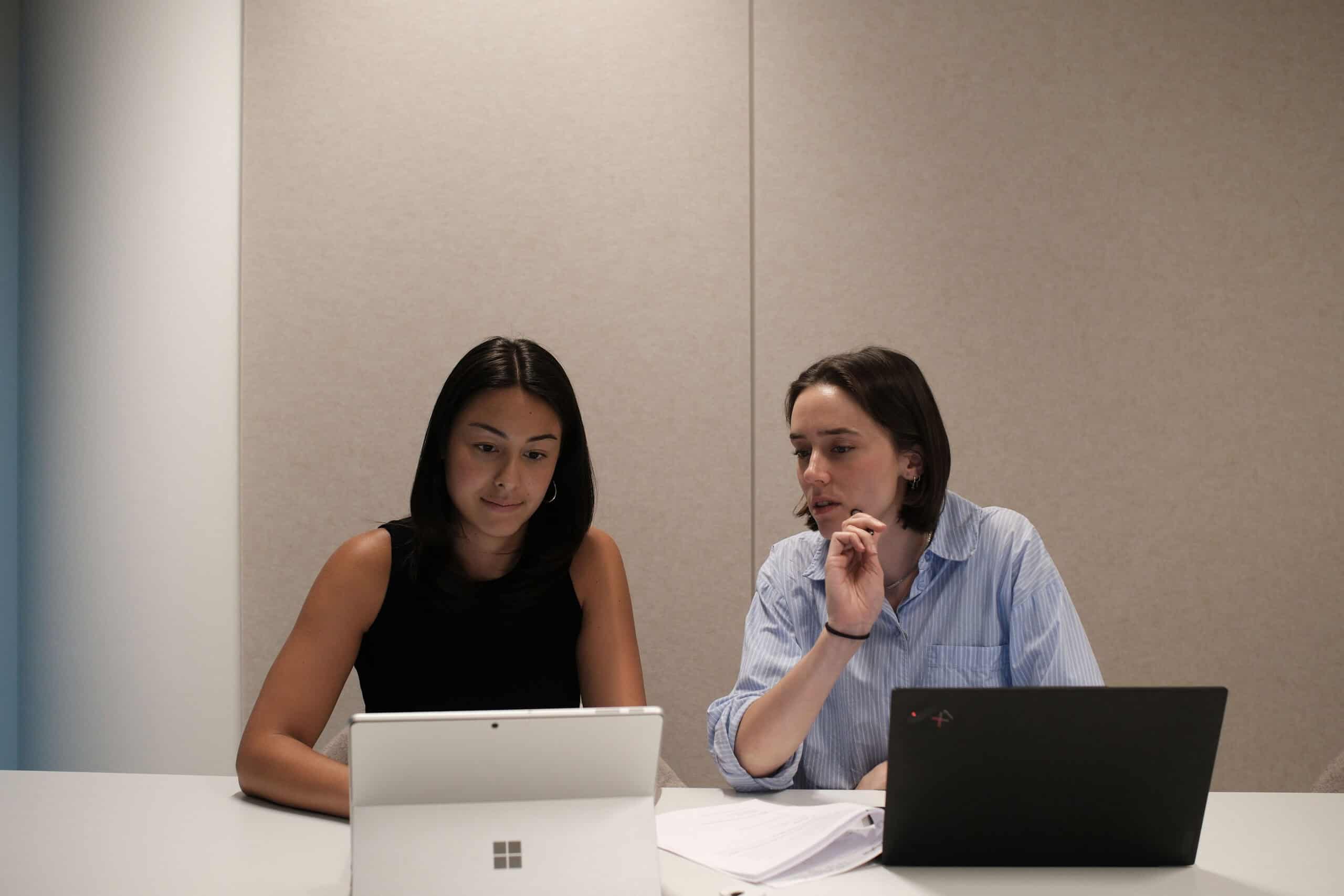 Two women working at a table with a tablet and a laptop. One looks at a screen, the other speaks.