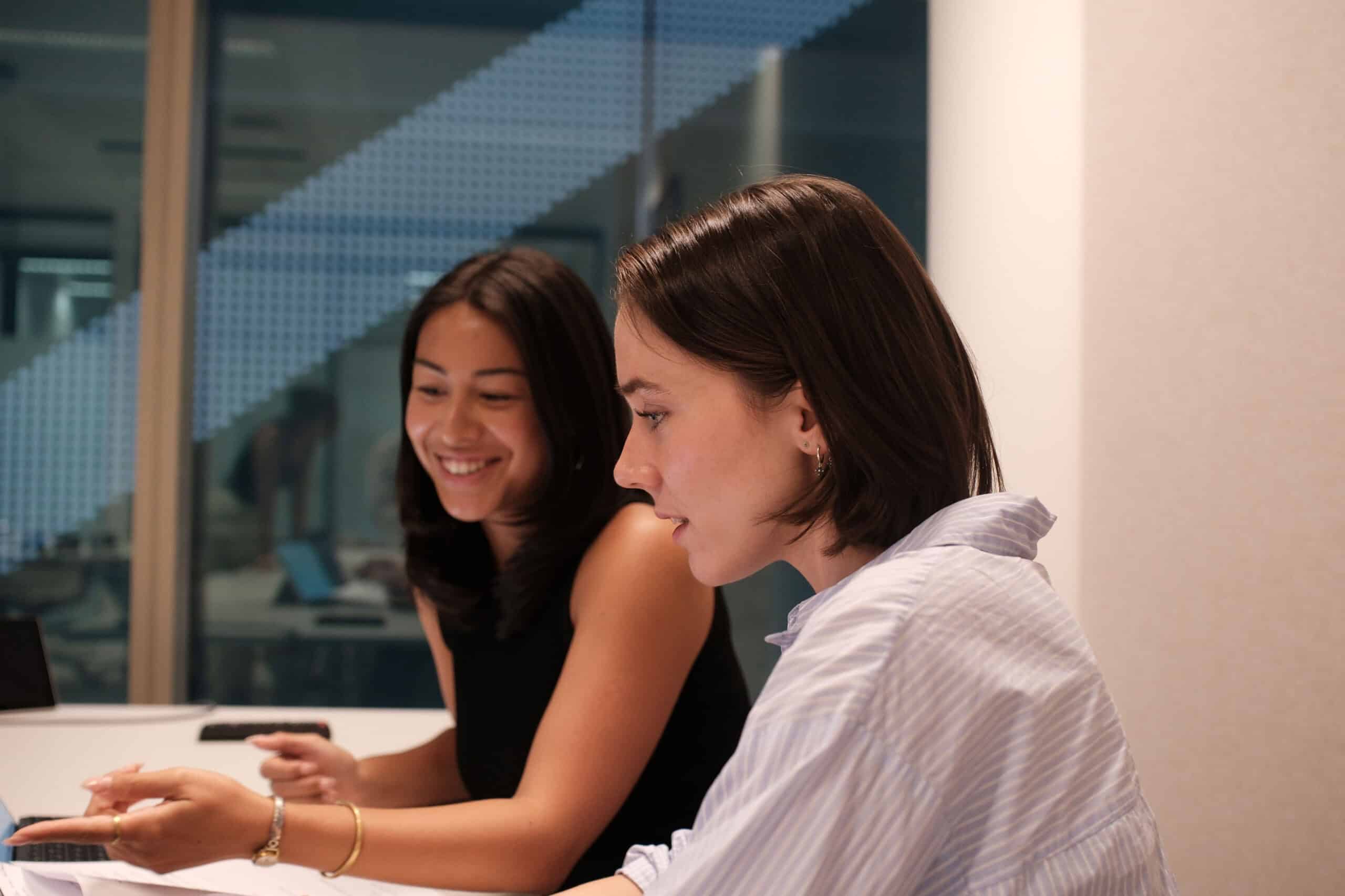 Two young women, one smiling and pointing, the other looking intently, discussing something at a desk in an office setting.
