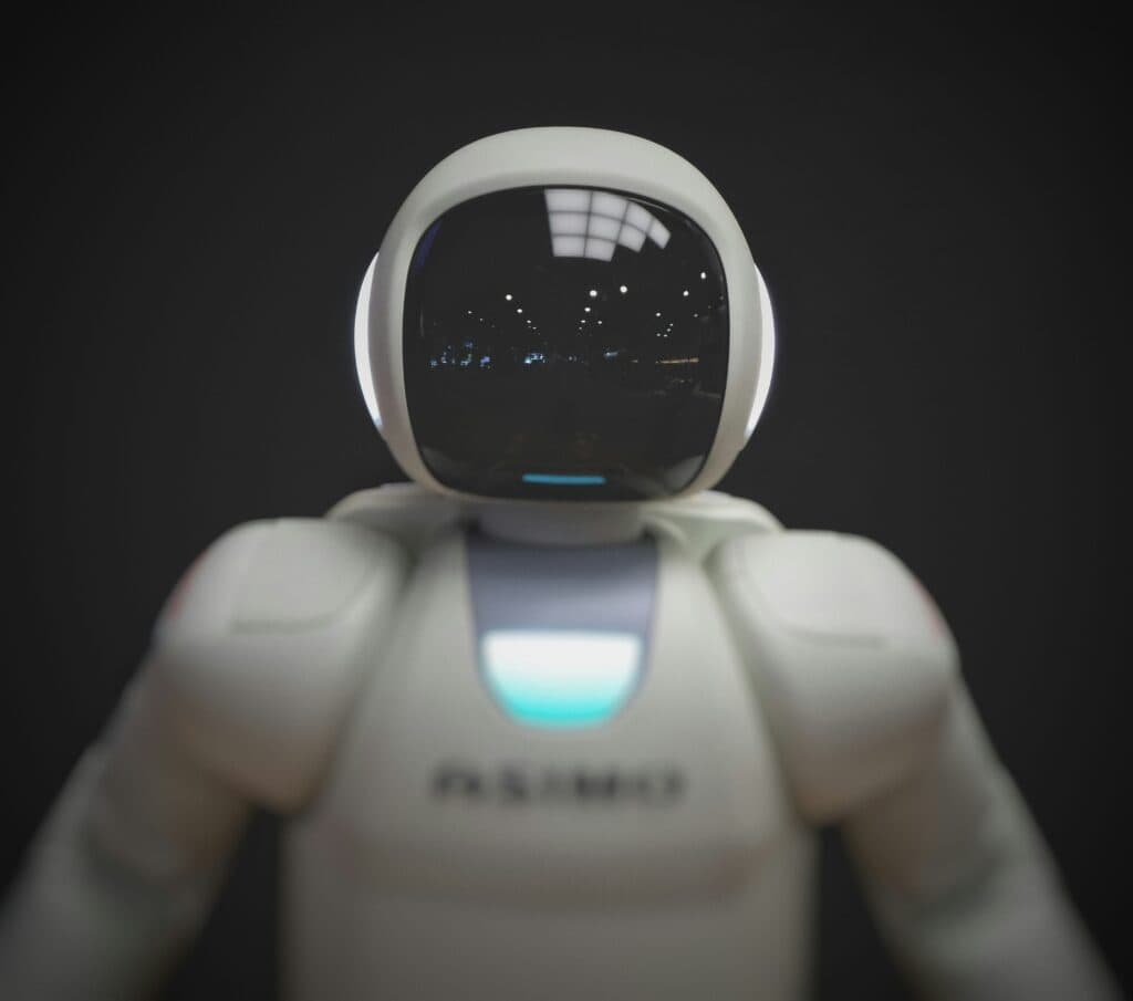 Close-up of a white robot with a dark, reflective visor showing light reflections, and a glowing blue chest panel.