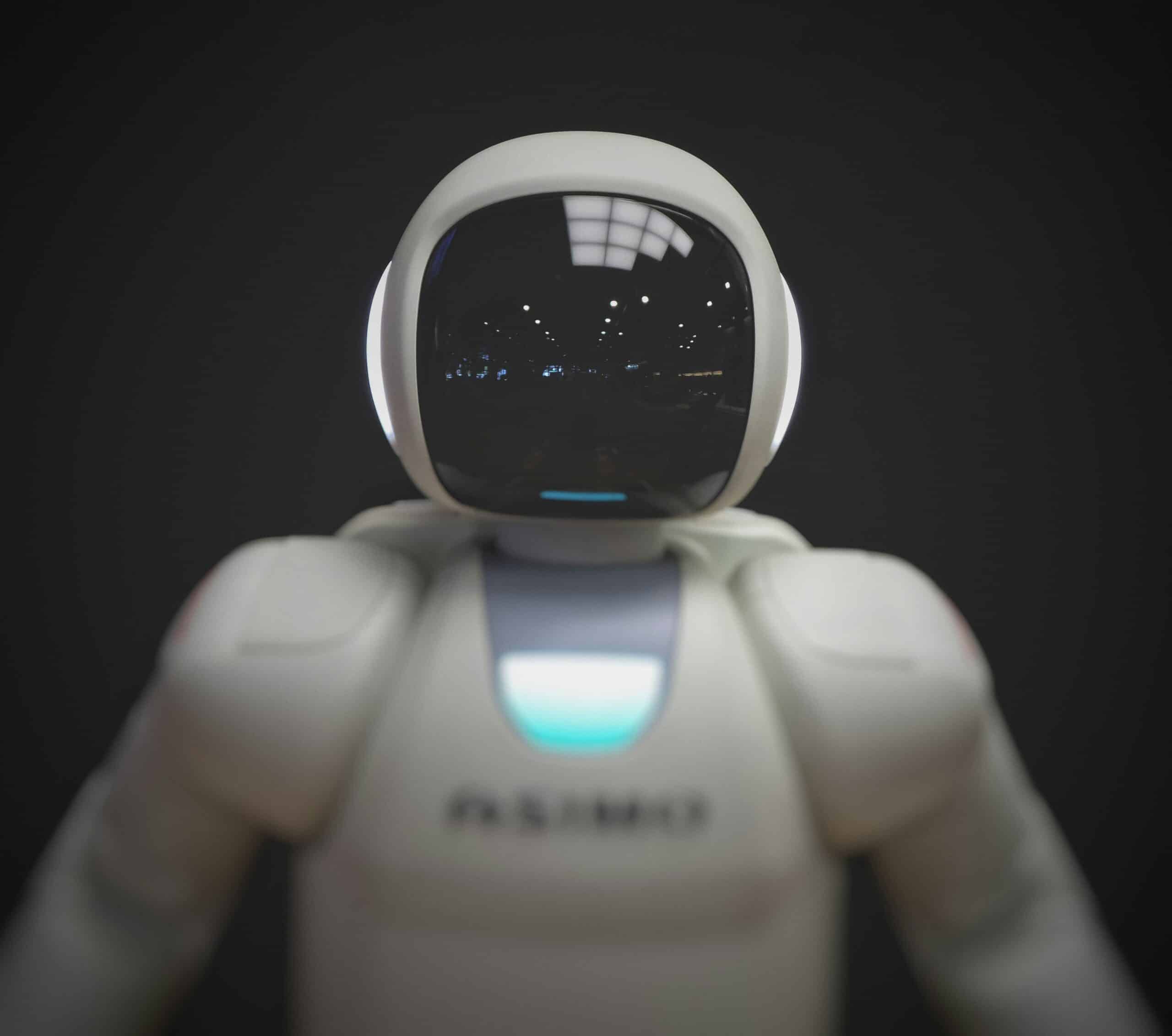 Close-up of a white robot with a dark, reflective visor showing light reflections, and a glowing blue chest panel.
