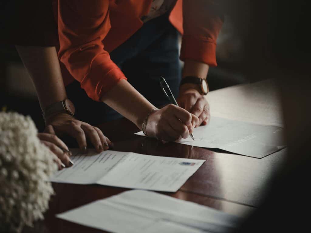 Close-up of two people's hands on a dark table, one signing a document with a pen.