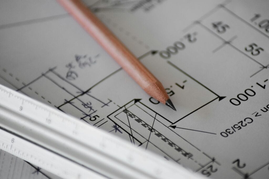 A sharpened wooden pencil and a white ruler resting on a blueprint with lines, numbers, and symbols.
