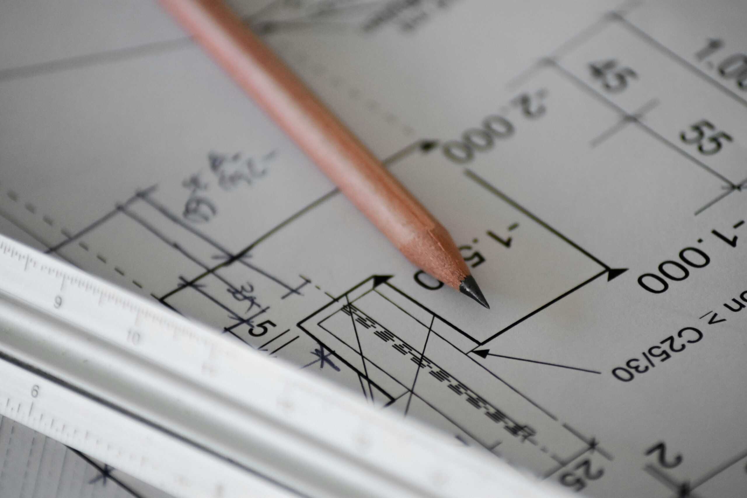 A sharpened wooden pencil and a white ruler resting on a blueprint with lines, numbers, and symbols.