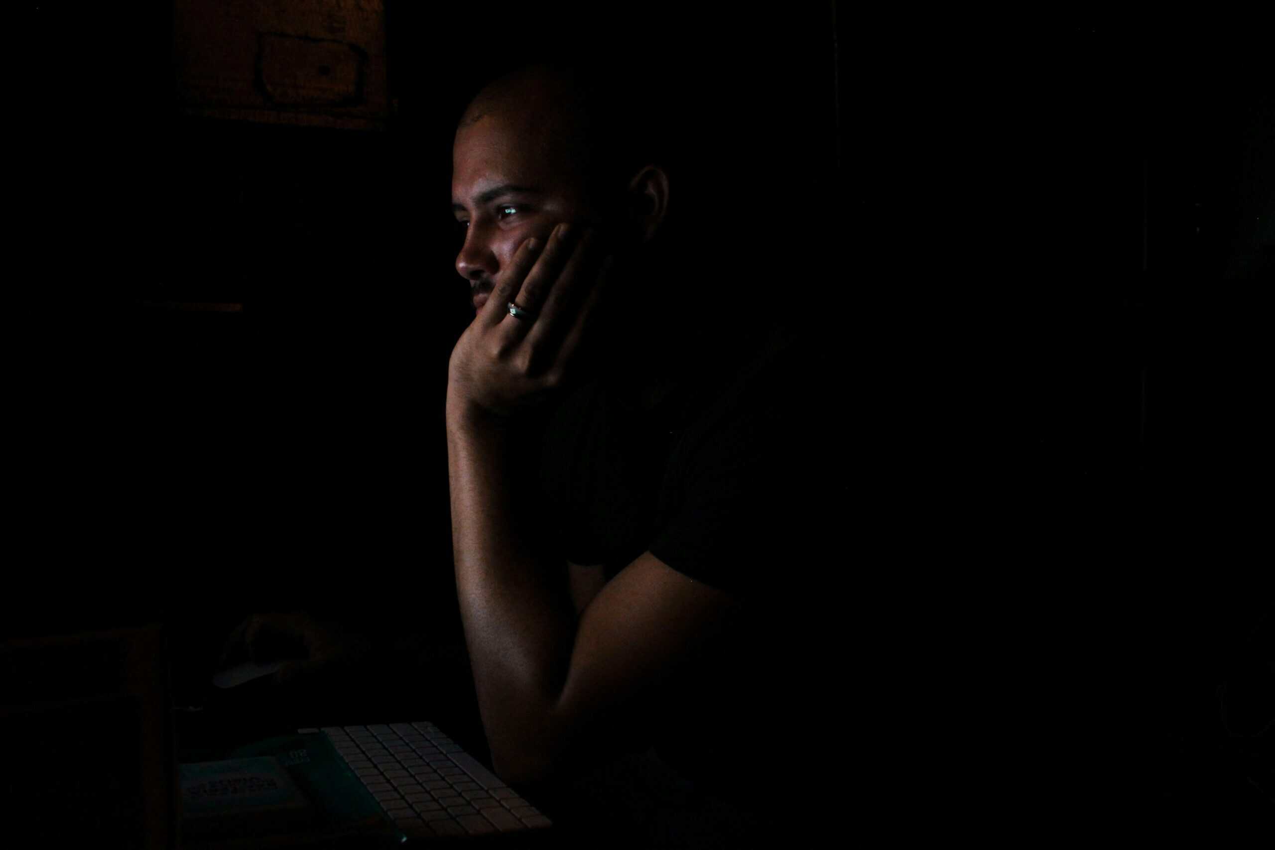 Side profile of a person with a shaved head and beard, hand on chin, looking intently at a screen in a dark room.