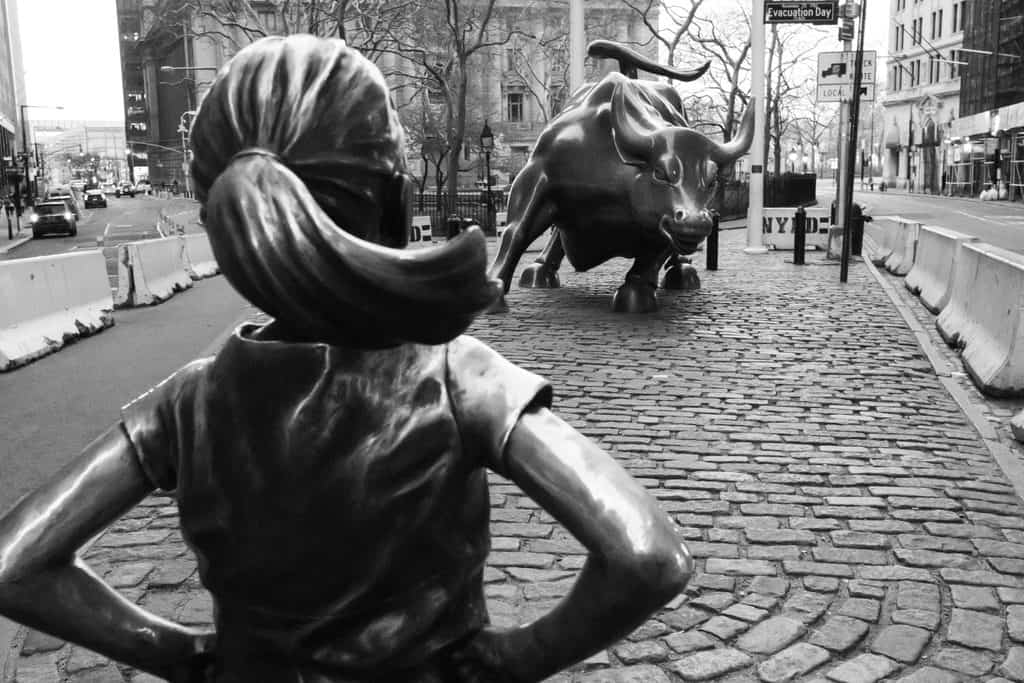 Black and white photo of the 'Fearless Girl' statue from behind, facing the 'Charging Bull' statue on a cobblestone street in a city.