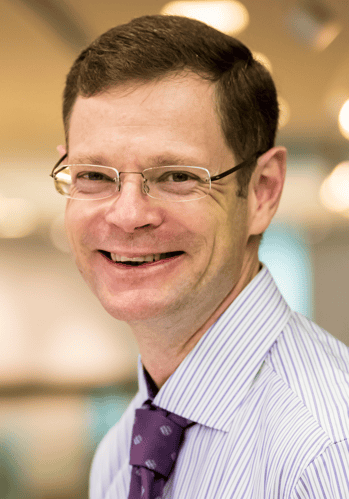 Headshot of a smiling man with glasses, wearing a striped shirt and purple tie.