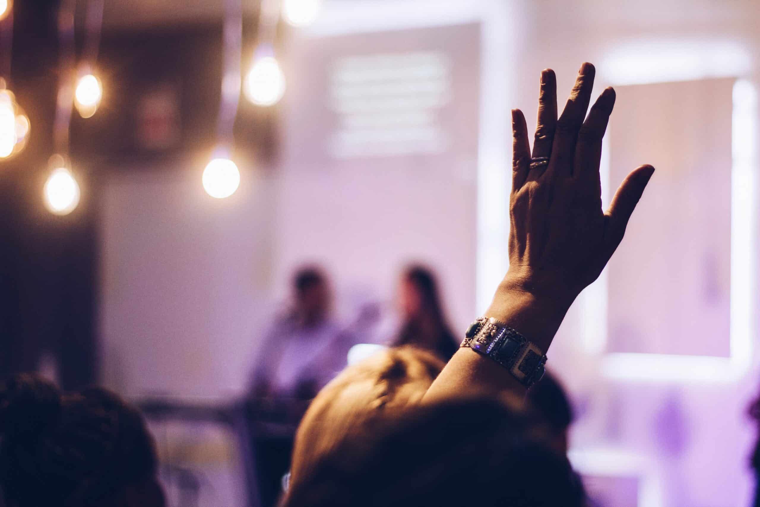 A person's right hand, wearing a ring and bracelet, raised in the air against a blurred background of lights and indistinct figures.