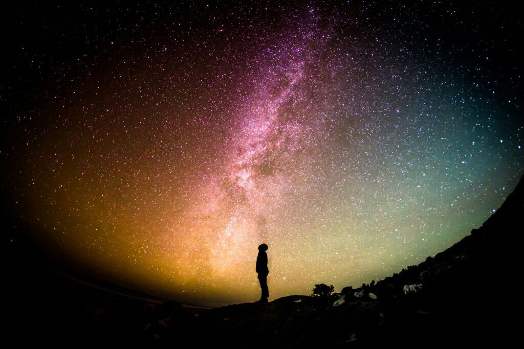 A person stands silhouetted against a colorful, star-filled night sky, looking up at the Milky Way which glows in shades of orange, pink, purple, and teal.