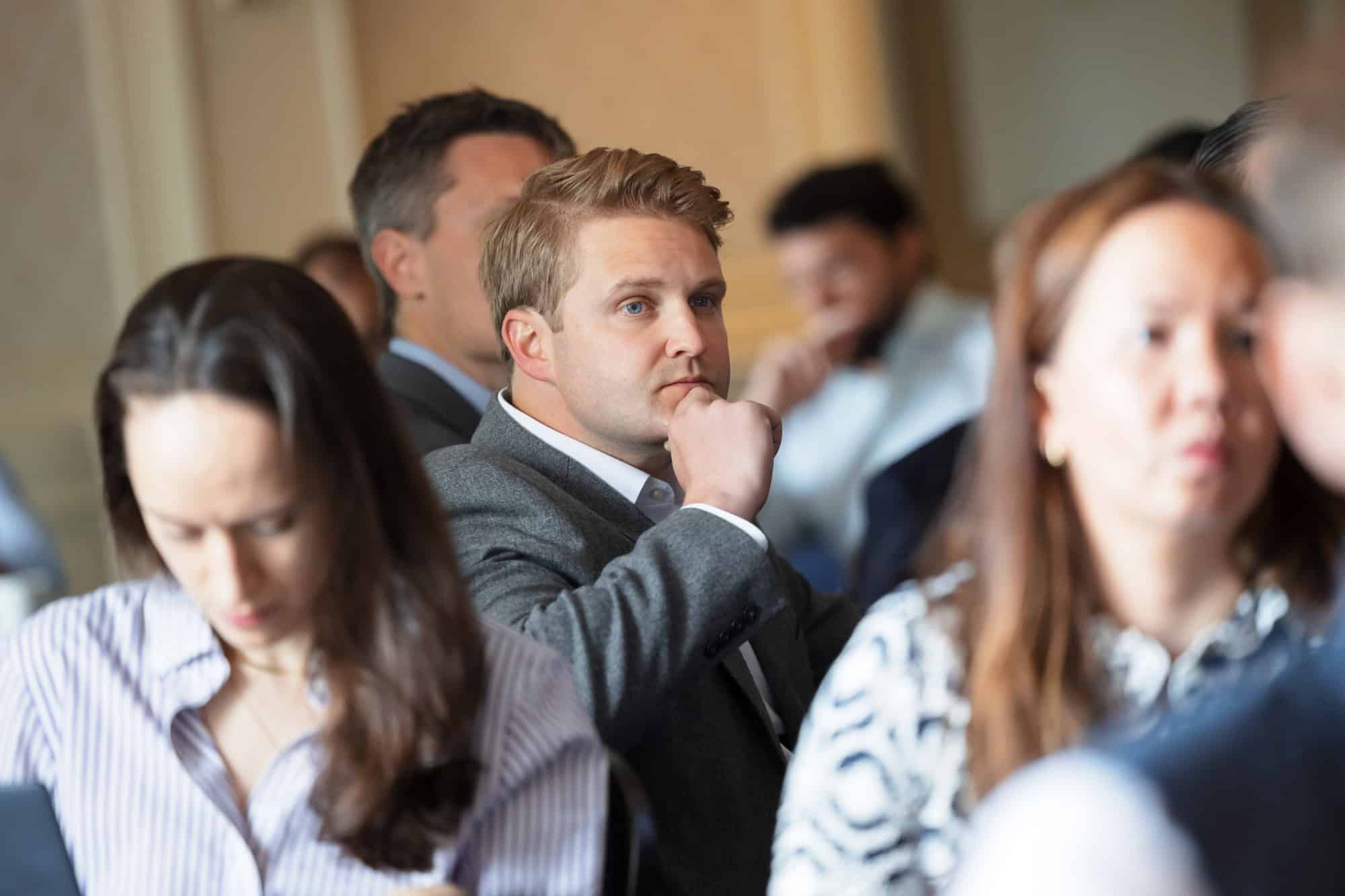 A man with blonde hair and blue eyes, wearing a grey suit, rests his hand on his chin while looking to the right. Other attendees are blurred in the foreground and background.