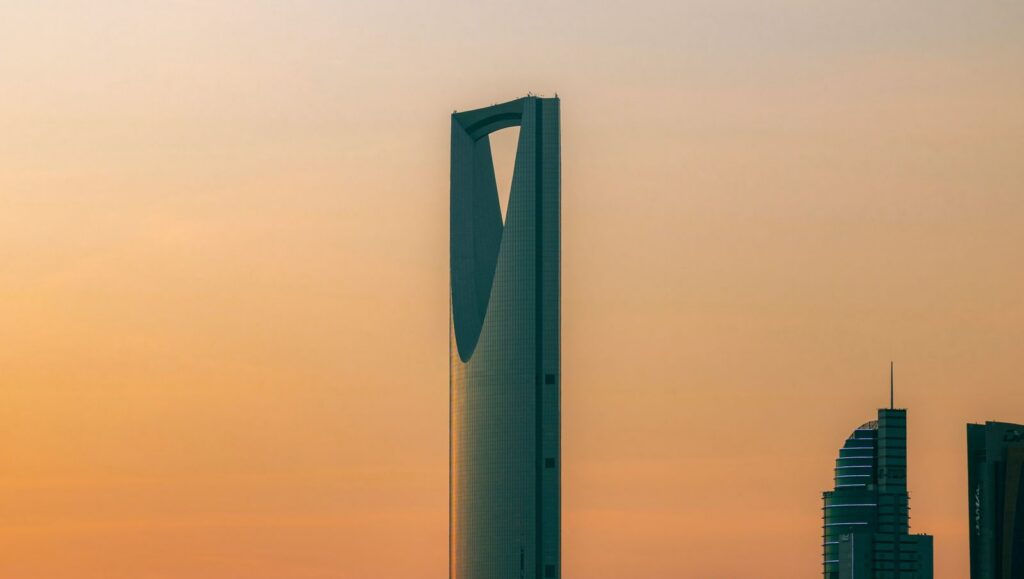 A tall, modern skyscraper with a distinctive inverted V-shaped opening at its top stands against a gradient orange and yellow sky, with other buildings and a construction crane visible to its right.